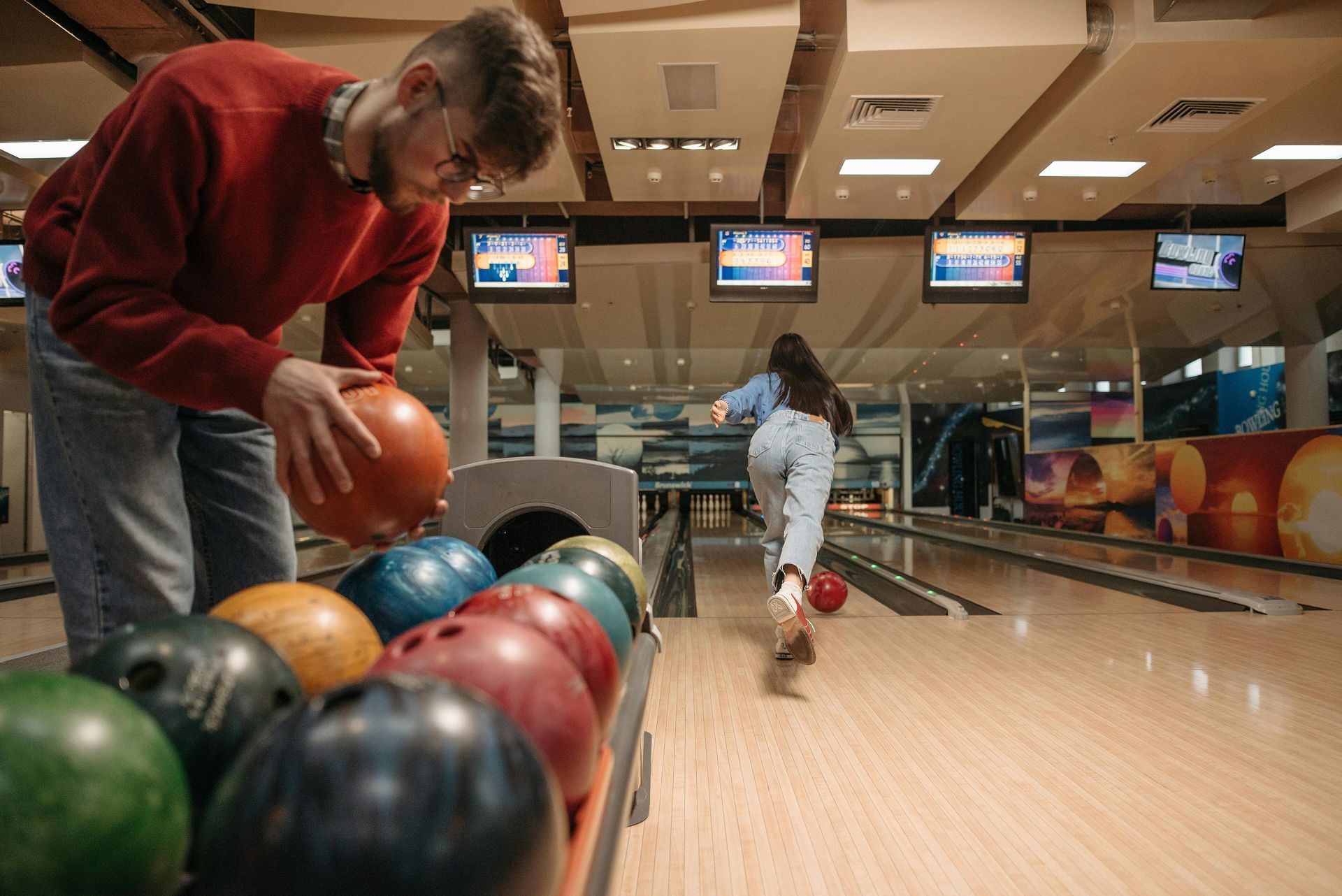 Man selecting a bowling ball; woman bowling. Bowling alley with multiple screens, balls in a rack.
