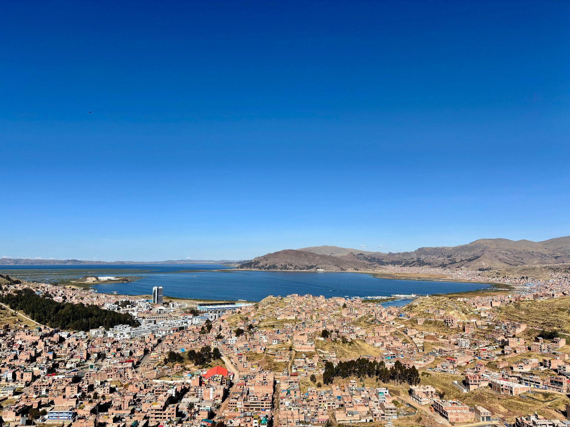 Vista aérea de Puno, Perú, con los edificios de la ciudad dominando las aguas del lago Titicaca bajo un cielo despejado.