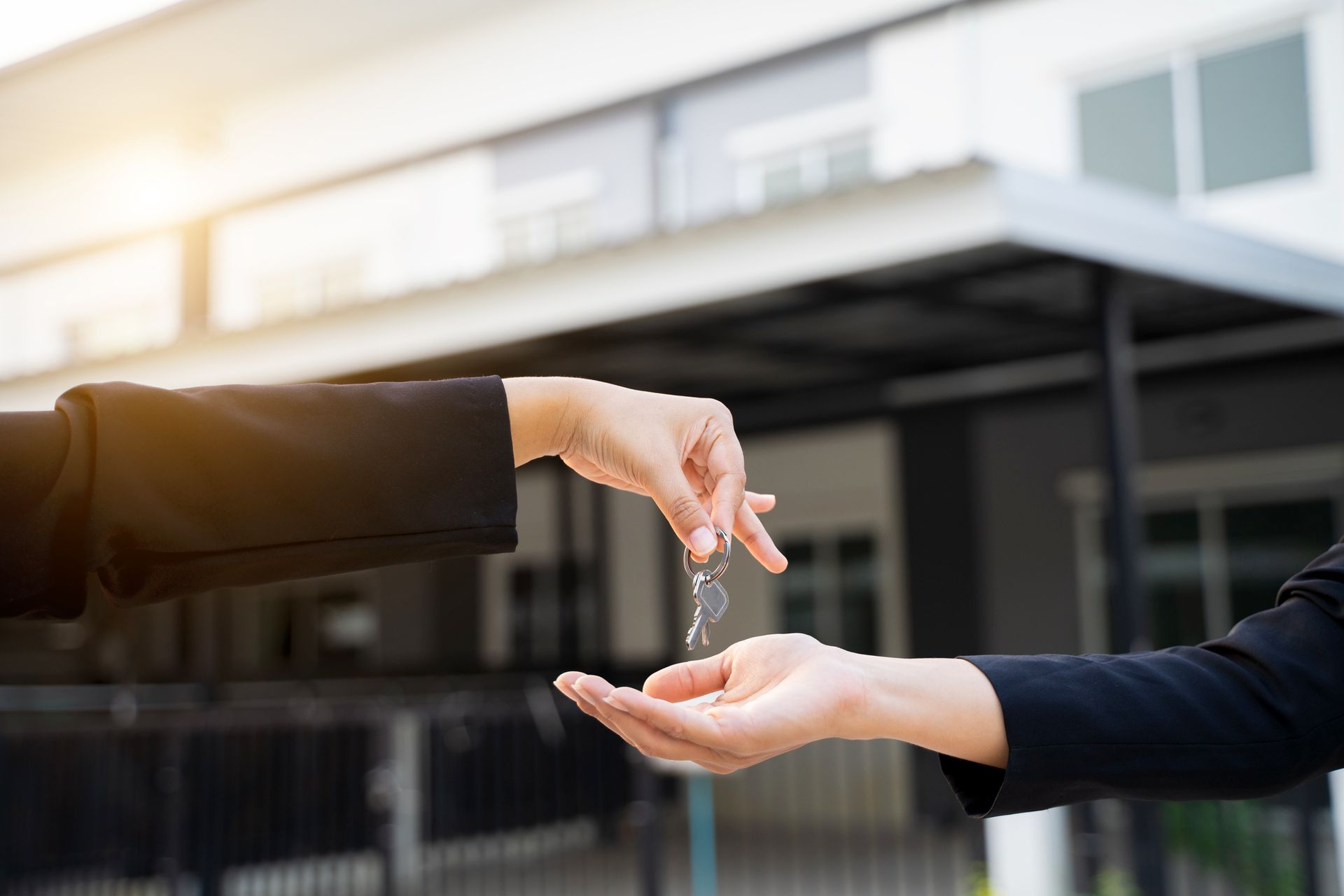 A person's hand offering keys to another person's hand, in front of a house.