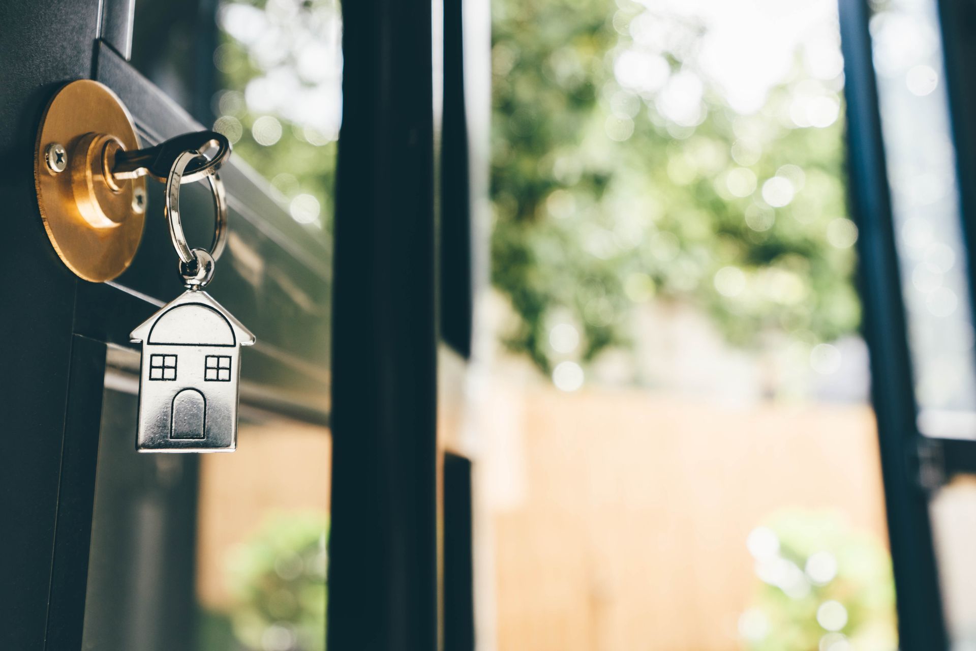 Key in door with house-shaped keychain; blurred outdoor view.