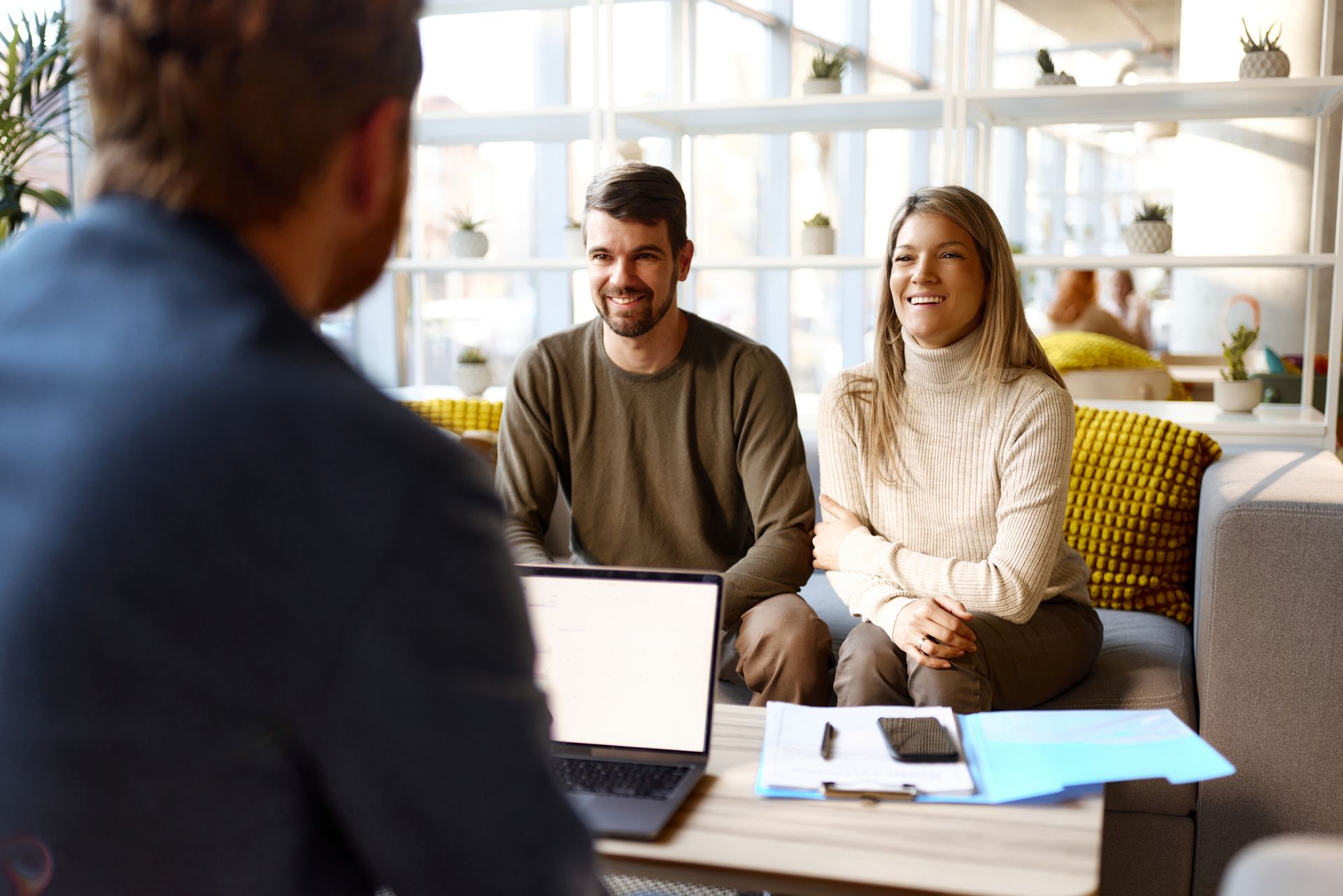 Man speaking to a smiling couple in a light-filled office. Laptop and papers on table.