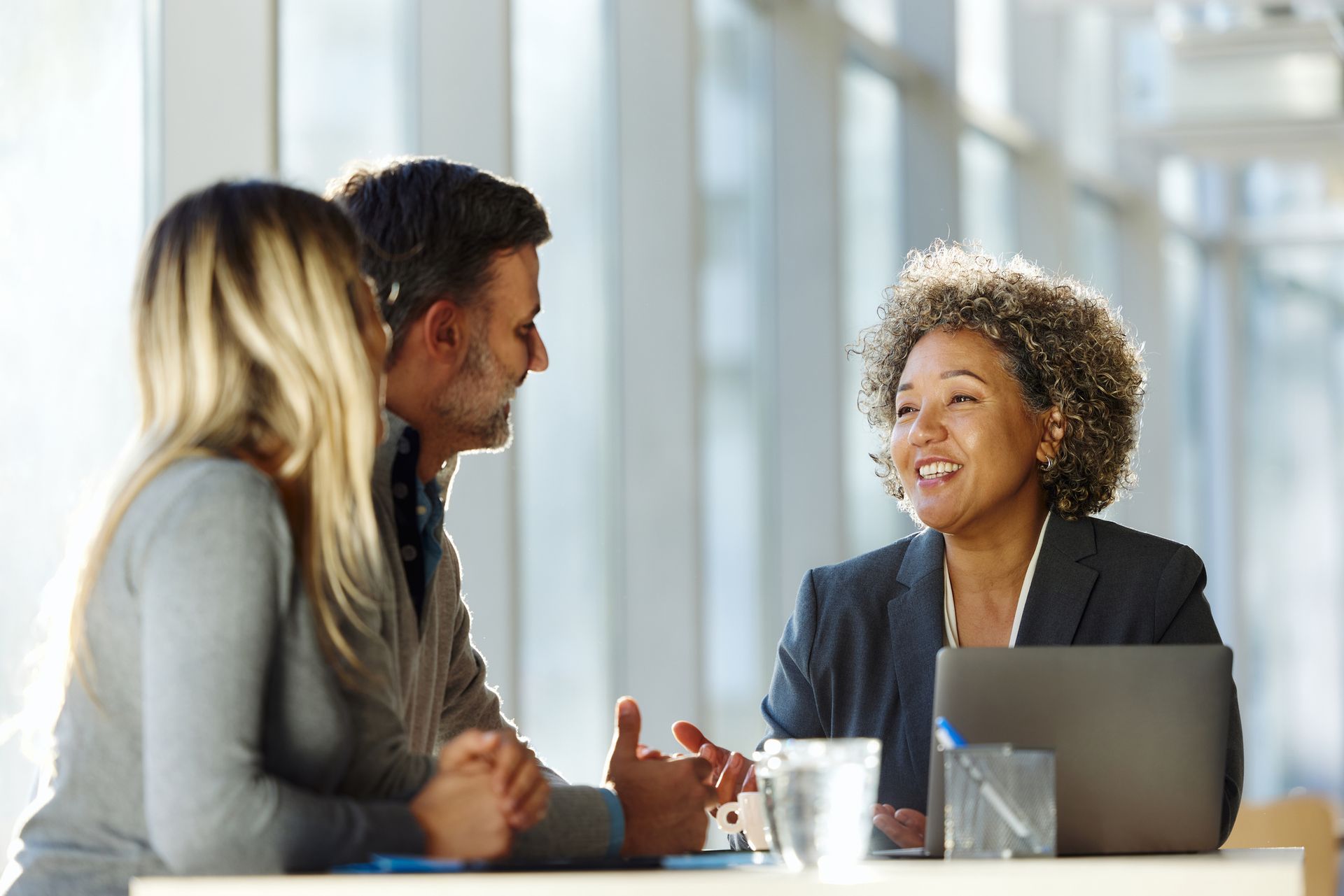 Woman smiling at a couple in an office setting, discussing over a laptop; sunlight.