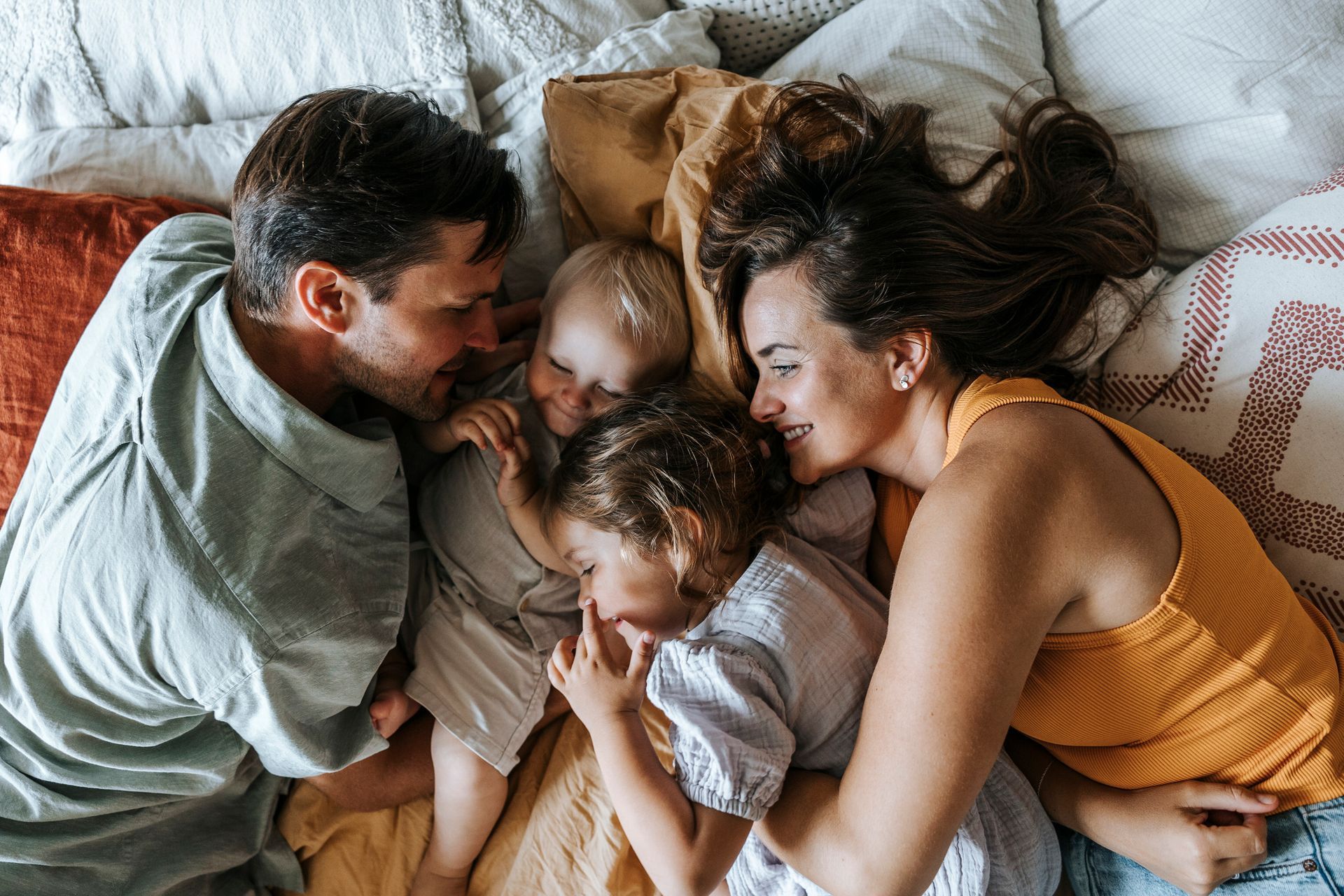 Family of four cuddling on a bed, smiling and looking at each other.