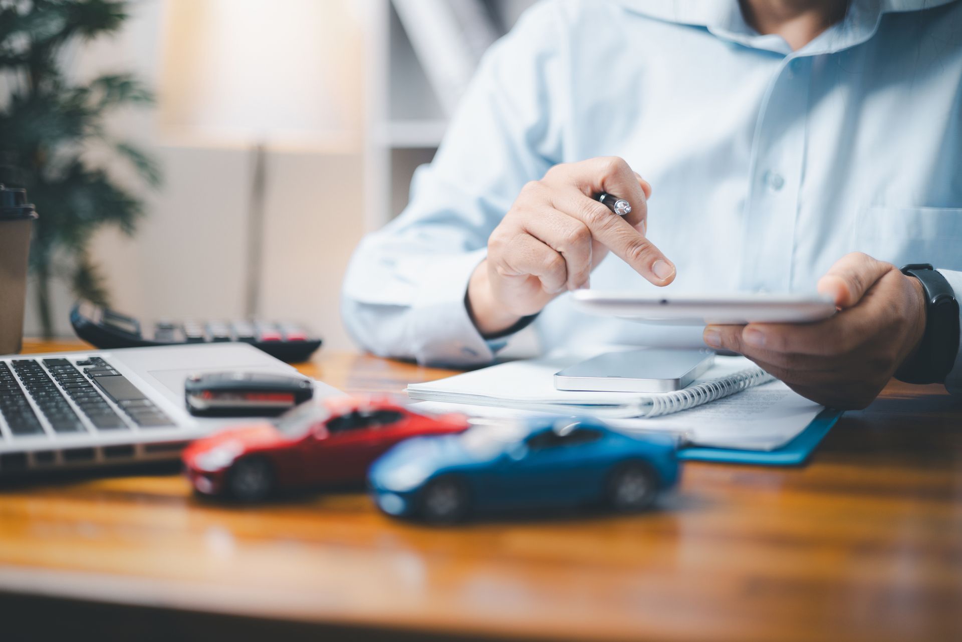 Person using a tablet with toy cars and a laptop on a desk.