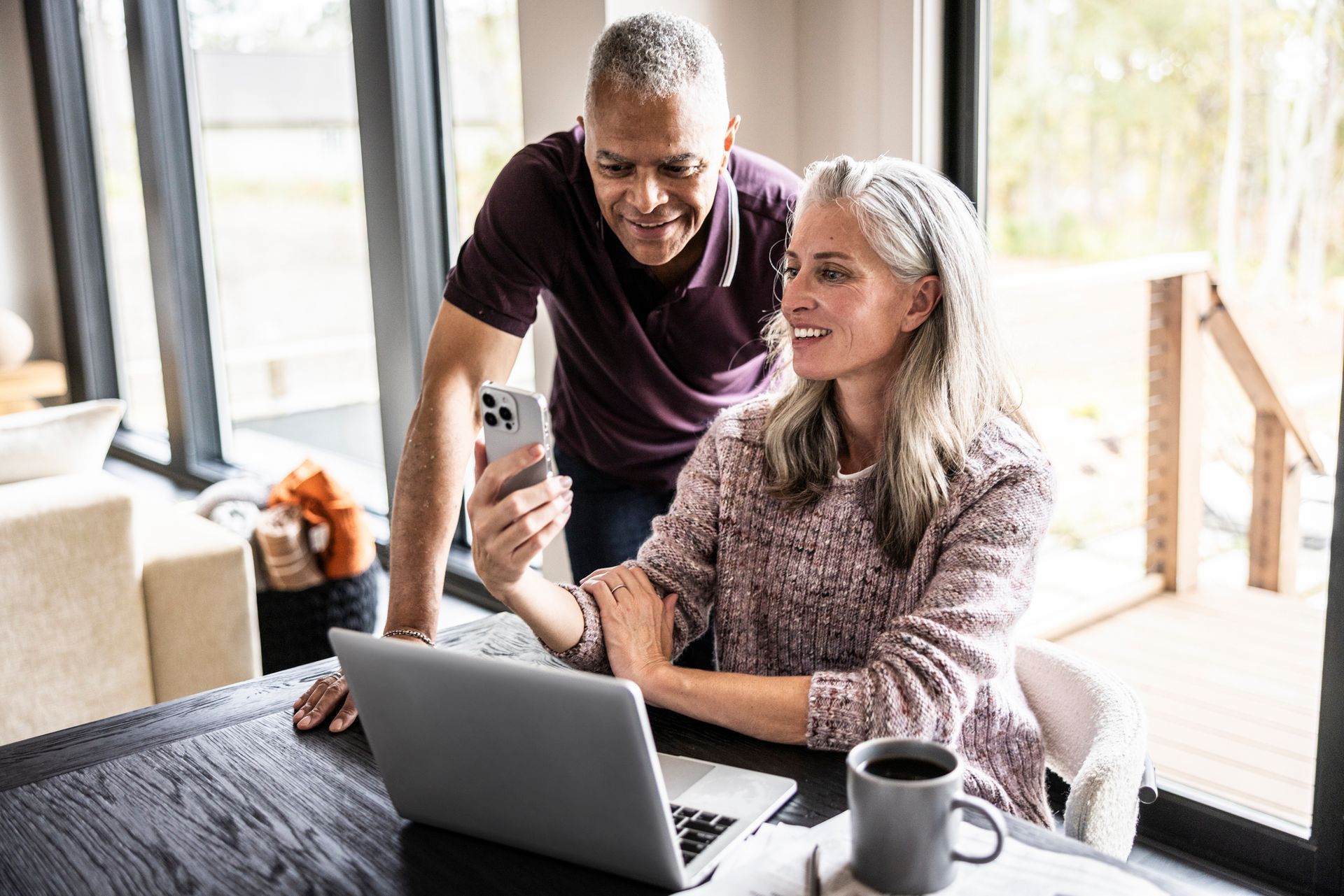 Couple looking at a phone, woman seated at a table with a laptop, coffee mug, sunny interior.