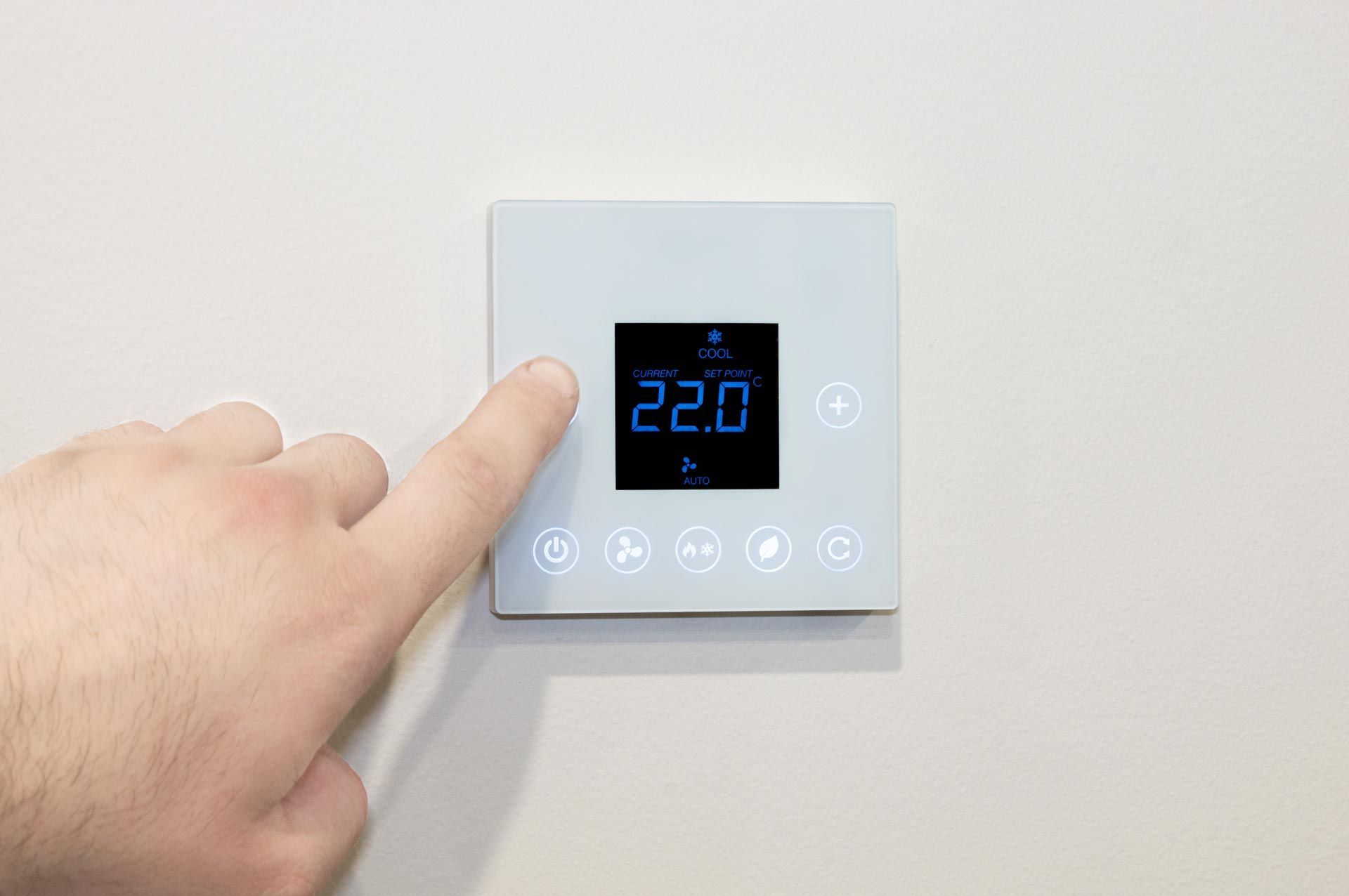 Hand pressing a button on a white digital thermostat displaying 22.0 degrees. Mounted on a light-colored wall