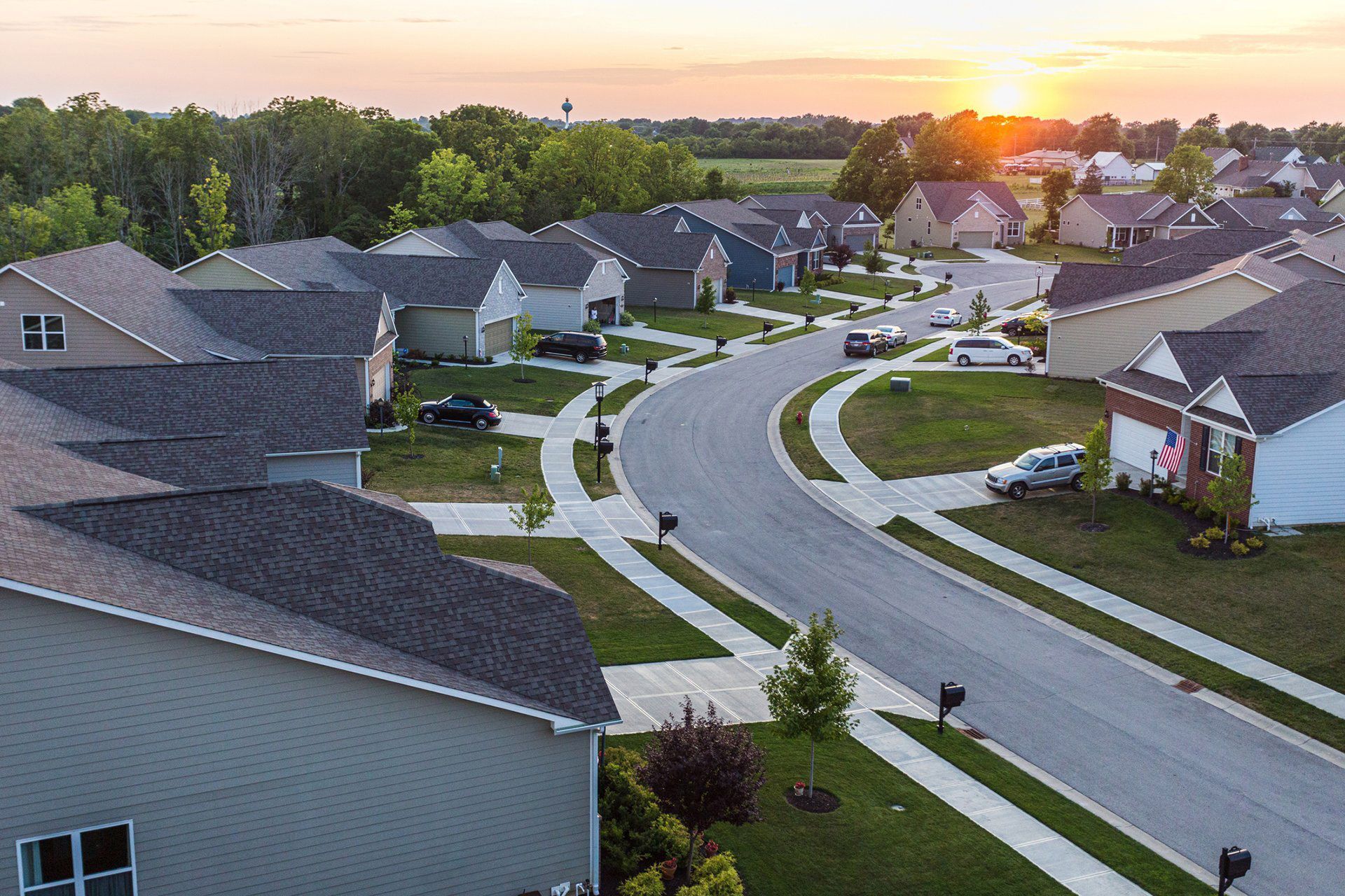 Suburban neighborhood with houses, curving road, green lawns, and sunset in the background.