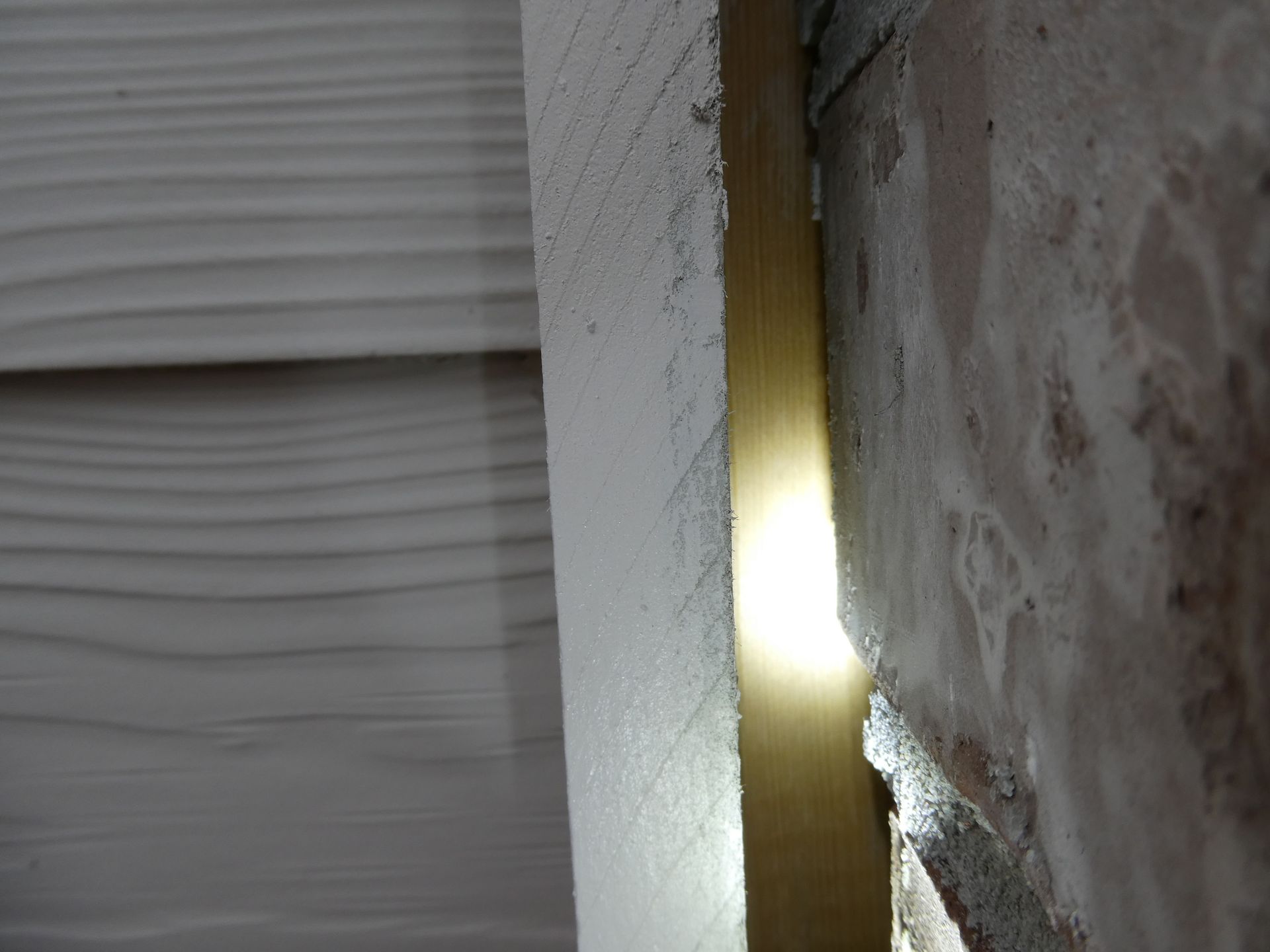 Close-up of a narrow gap between white siding and a rough brick wall, with light shining through the opening.