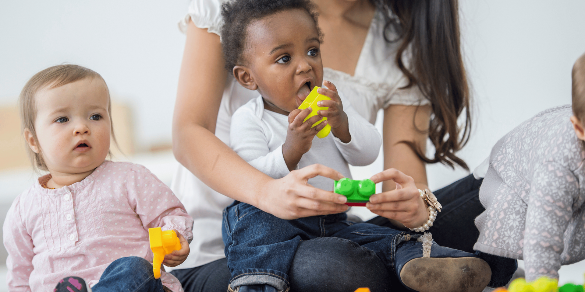 Babies and caregiver playing with blocks. Baby in caregiver's lap has a block in its mouth.