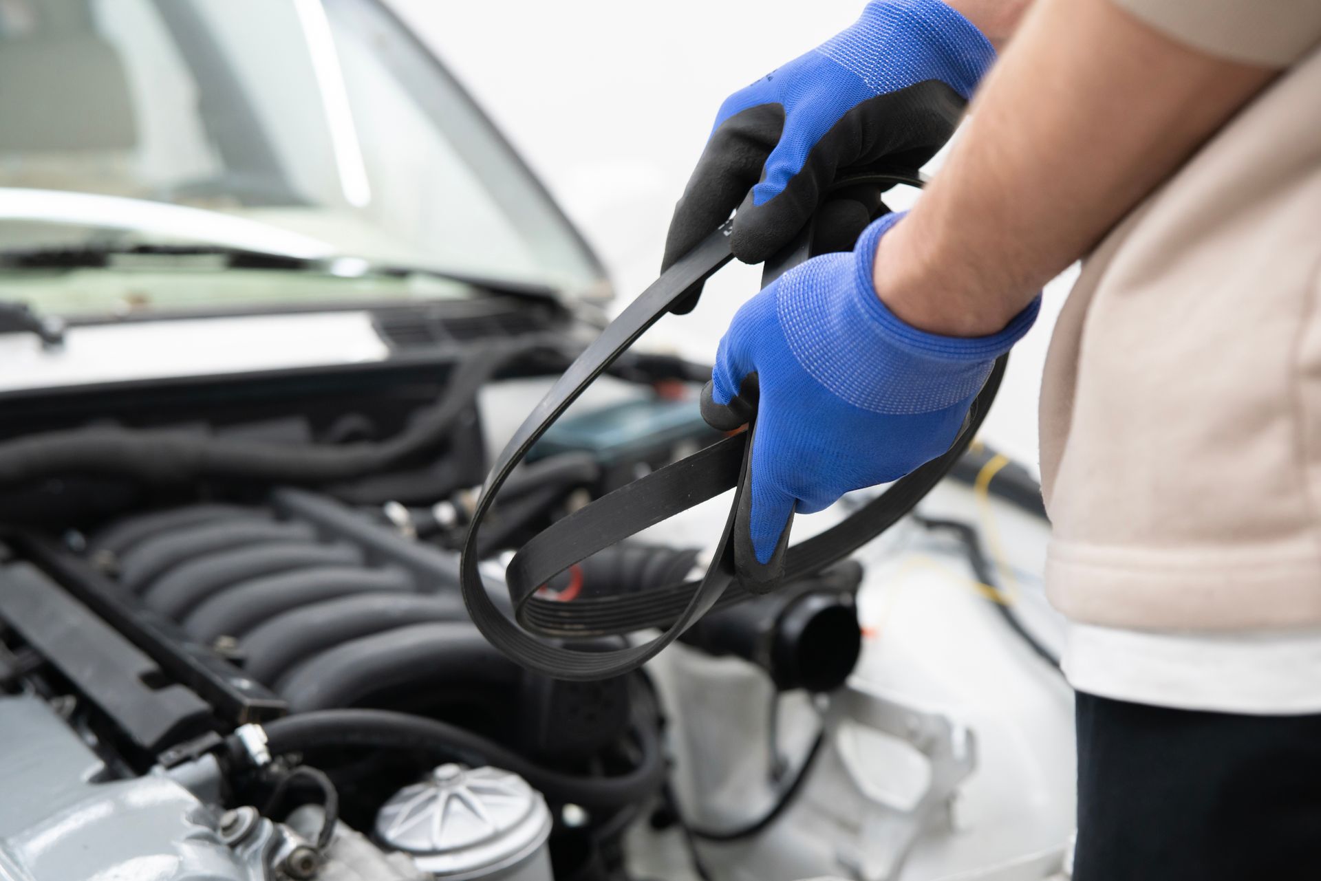 Mechanic holding a black serpentine belt near a car engine, wearing blue gloves.