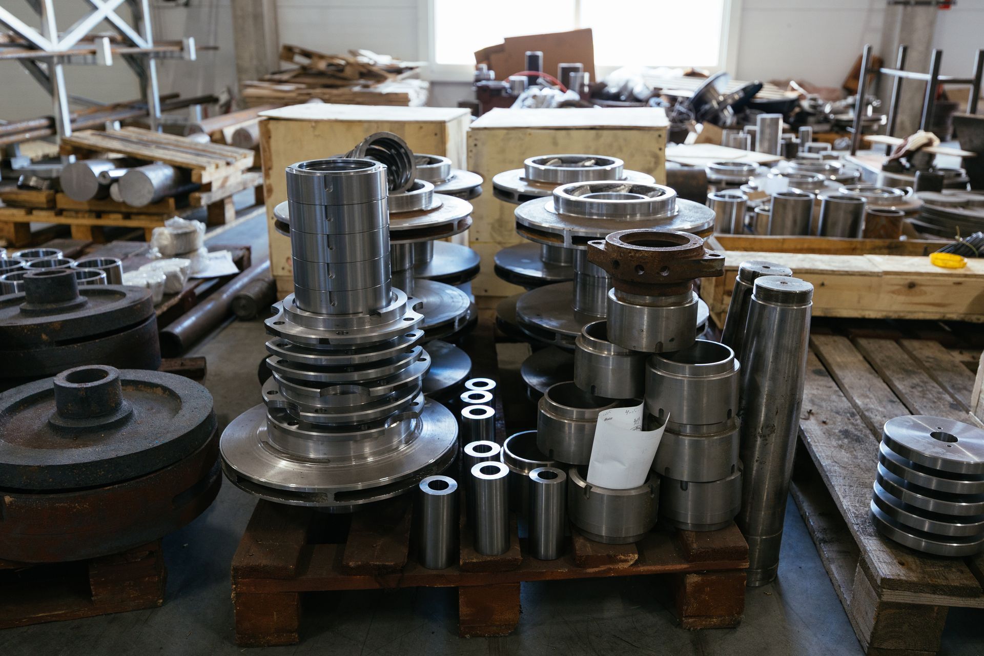 Metal machine parts on pallets in a warehouse, including discs, cylinders, and housings.