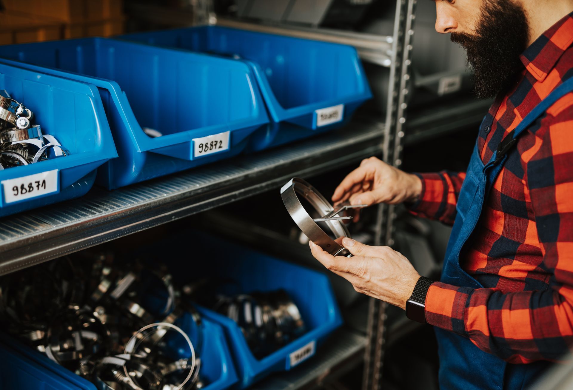 Person in a red plaid shirt inspects metal parts in a warehouse with blue storage bins.