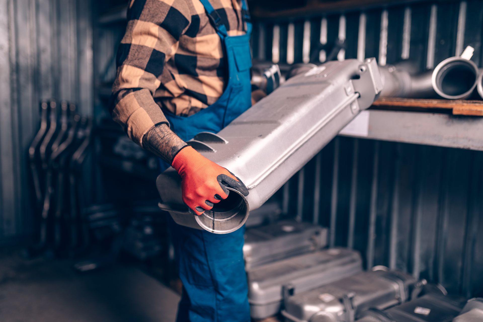 Mechanic holding a silver exhaust part in a storage room; red gloves, plaid shirt, and blue overalls.