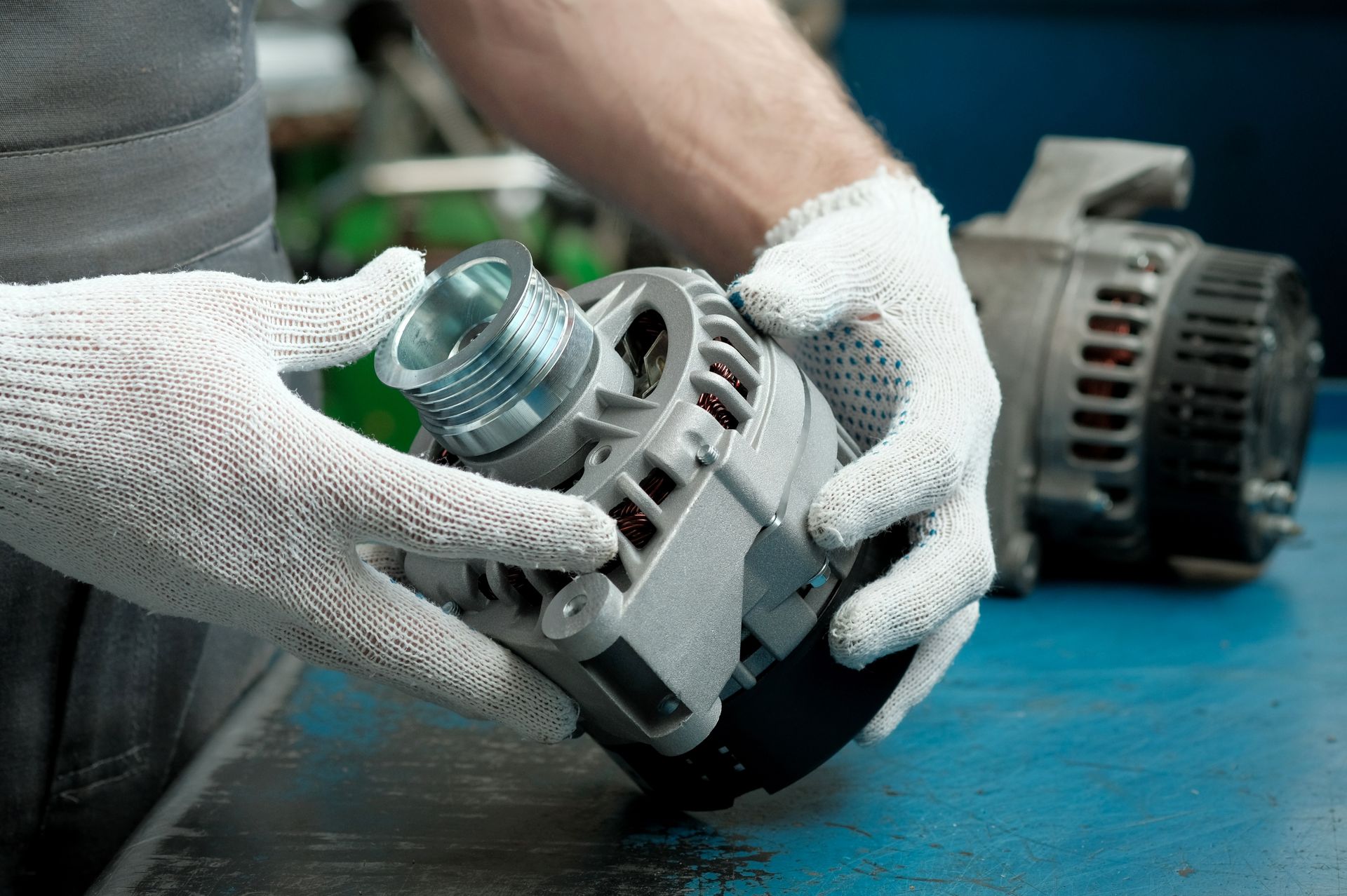 Mechanic in gloves holding a car alternator; another alternator sits nearby.