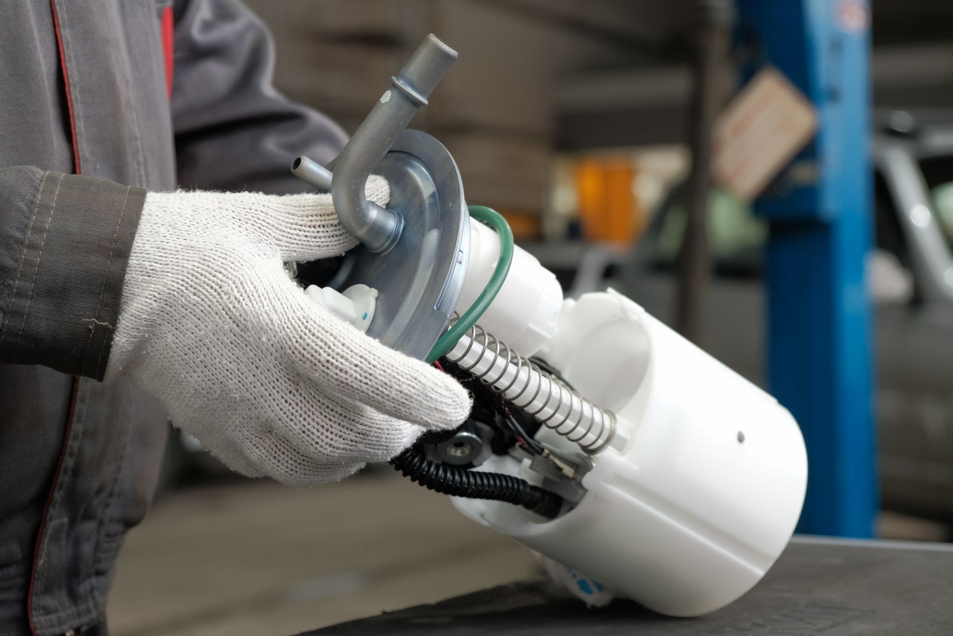 Mechanic's gloved hands holding a car fuel pump assembly in a garage setting.