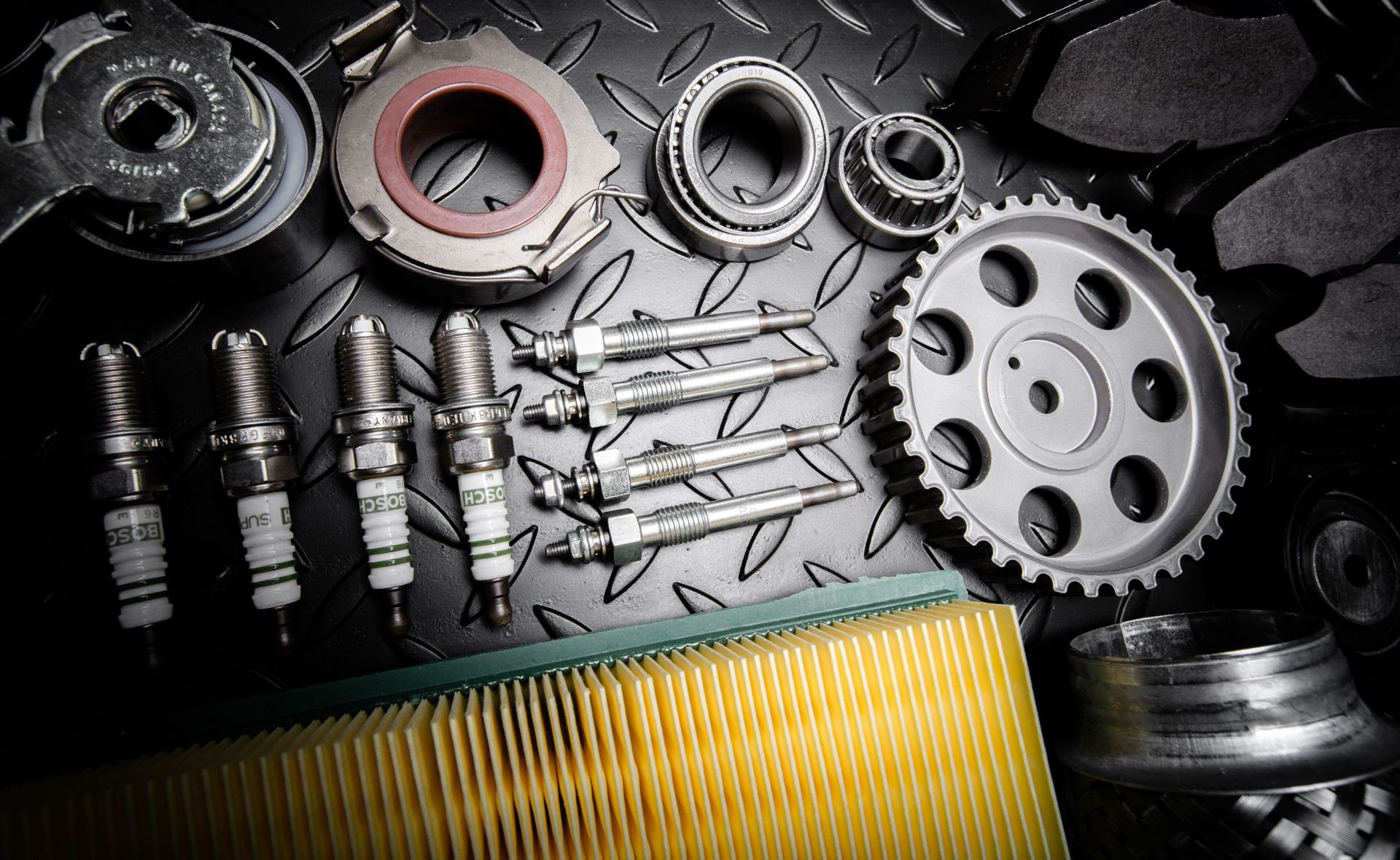 Car parts arranged on a black diamond plate surface: spark plugs, bearings, filters, and gears.