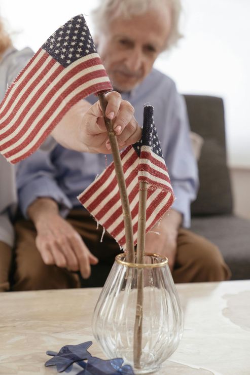 An elderly man is holding two small american flags in a vase.
