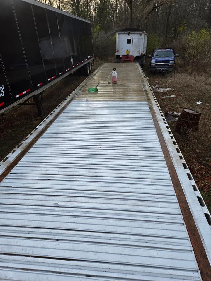 Flatbed trailer being washed, with soap and brush. Trailer is parked near a small building and truck, in an outdoor setting.