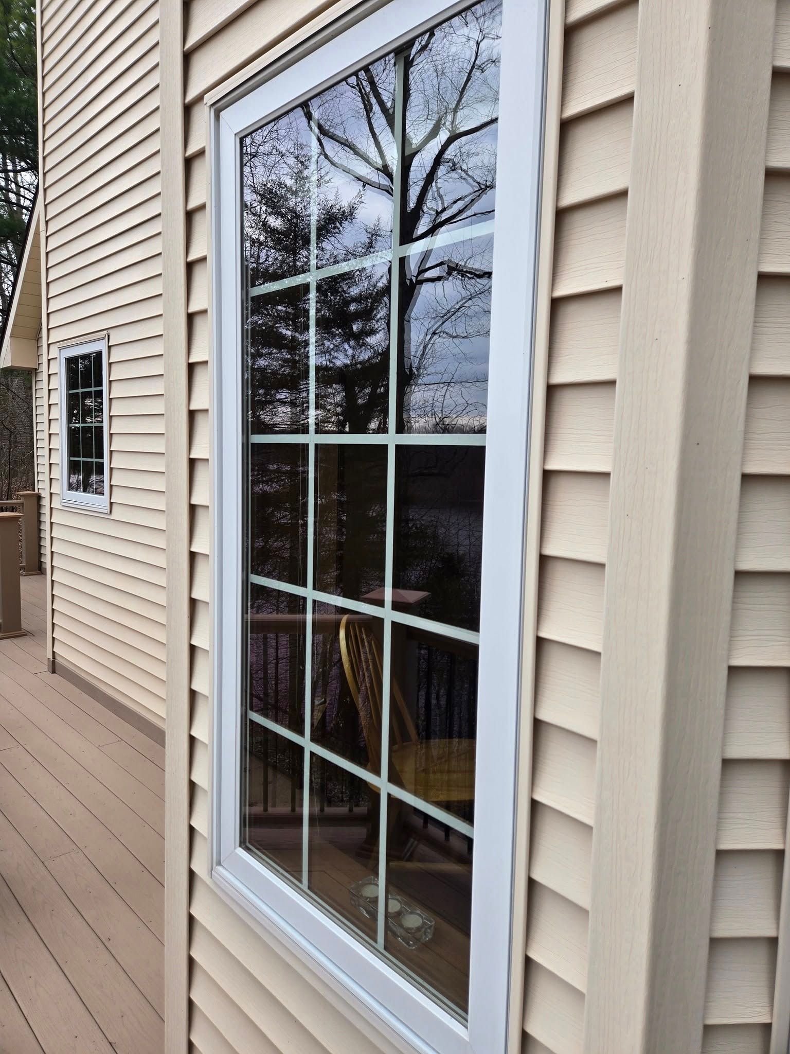 Tall, white-framed window on a tan-sided house with a grid pattern. Reflects trees and a deck.