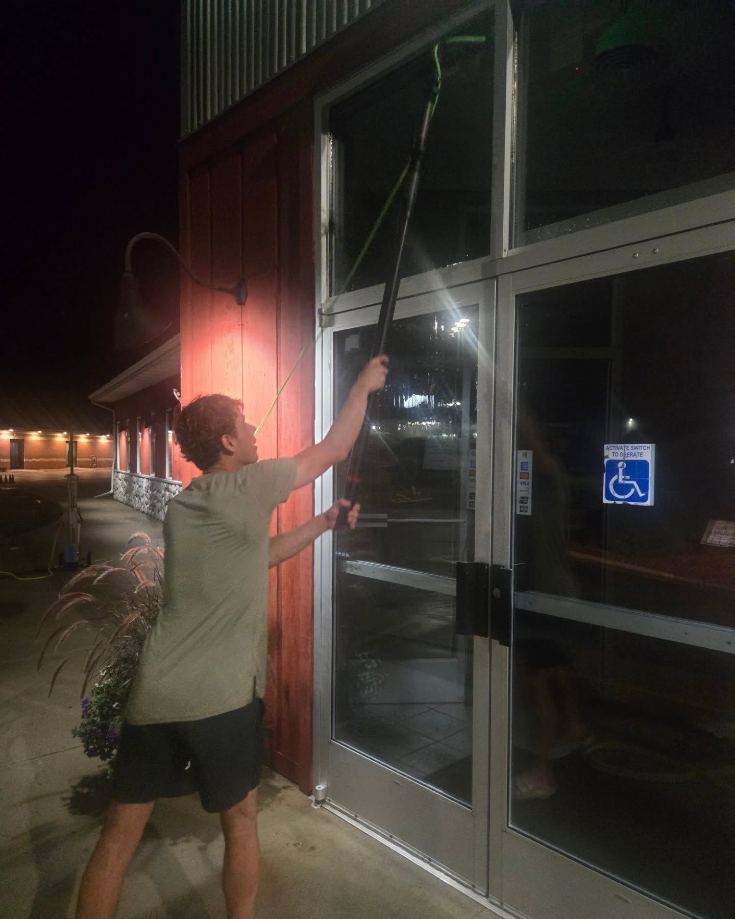 Person washes a window with a long-handled tool at night. Red building exterior with a handicapped parking sign visible.