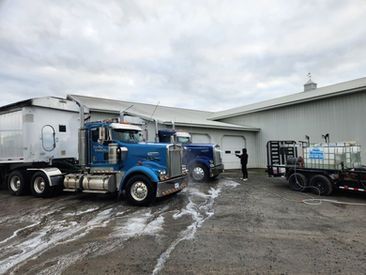 Blue semi-trucks being washed outside a large building. A person hoses down one truck, the pavement wet with suds.