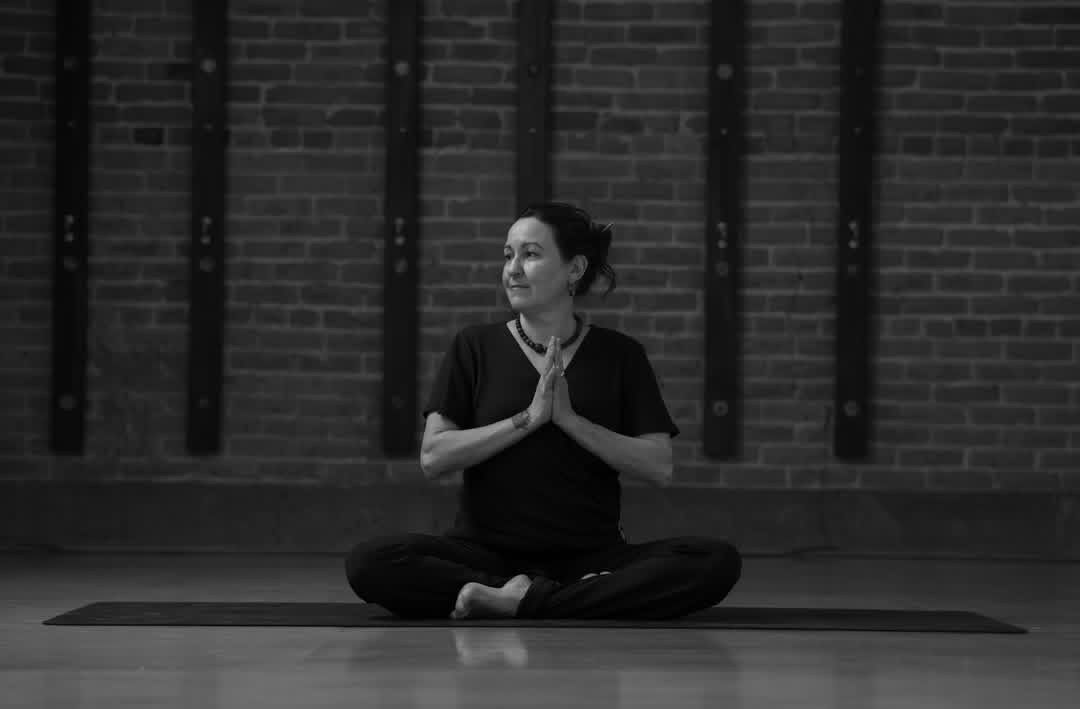 A woman is standing on one leg on a yoga mat in front of a brick wall.