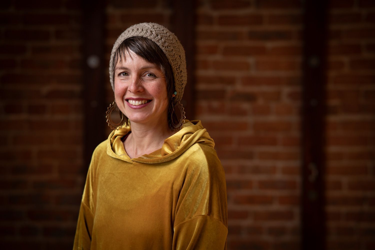 A woman wearing a hat and a yellow hoodie is smiling in front of a brick wall.