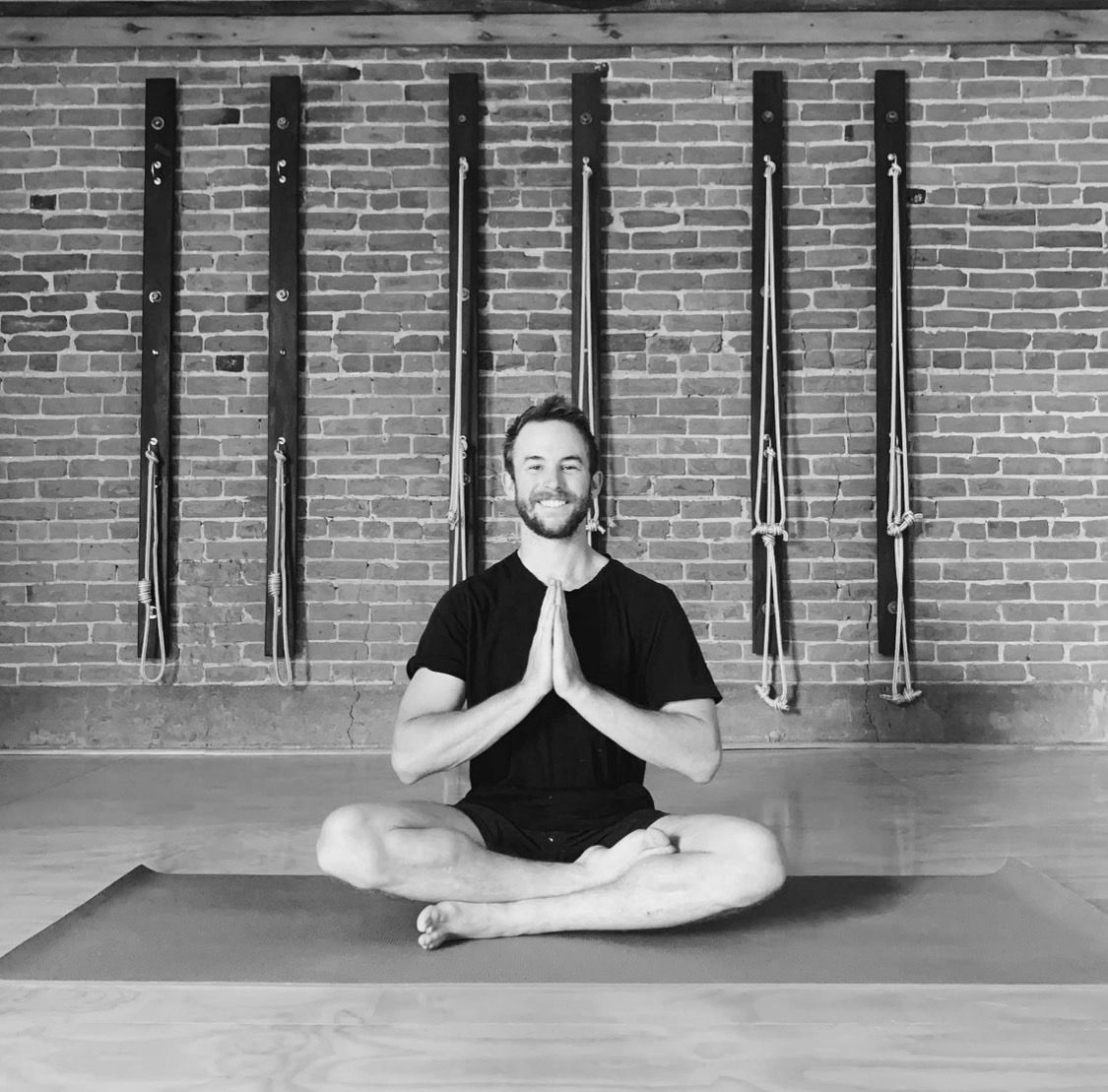 A man is sitting in a lotus position on a yoga mat in front of a brick wall.