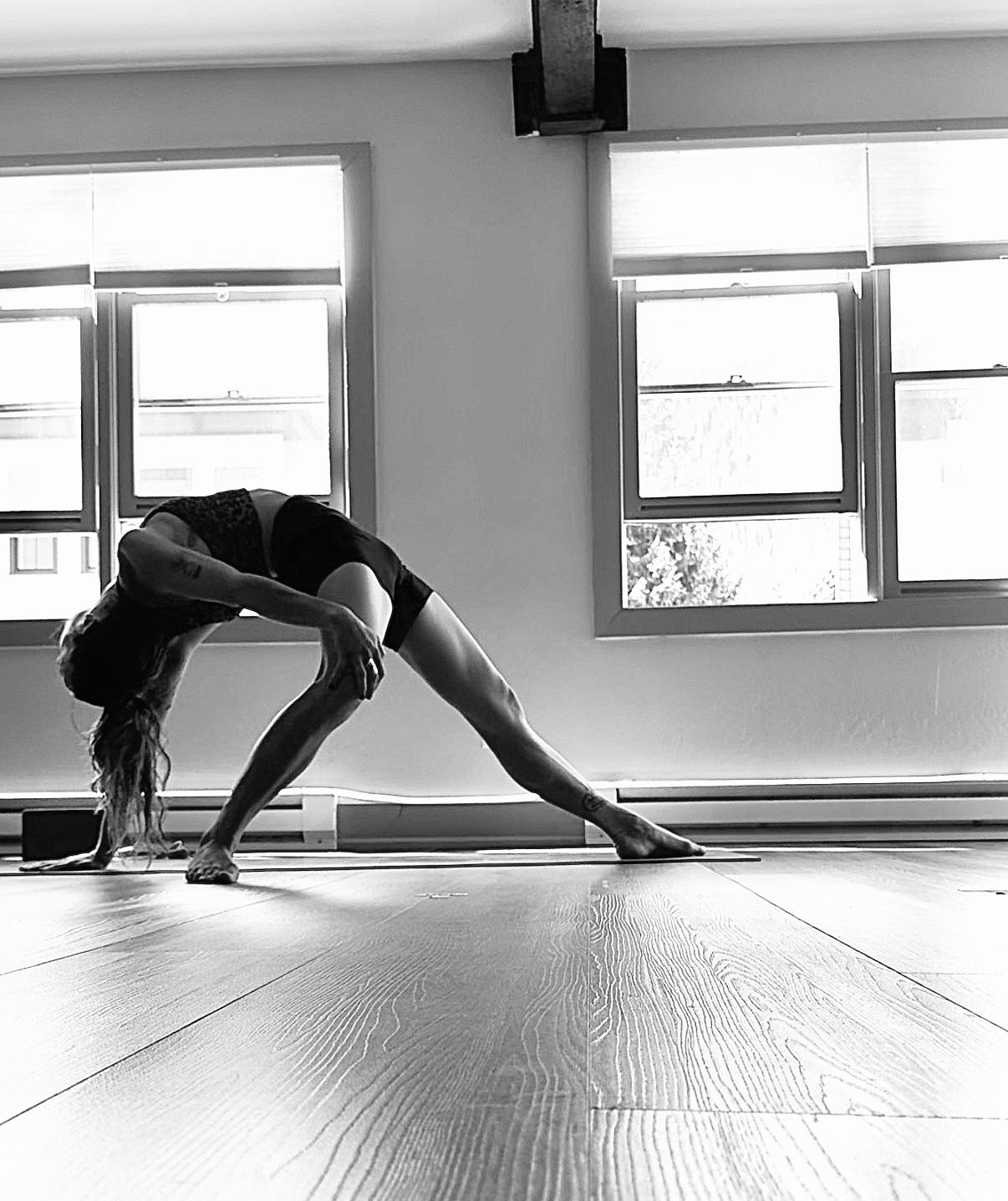 Woman in downward-facing dog pose with one leg extended upwards, on yoga mat. Black and white photo.