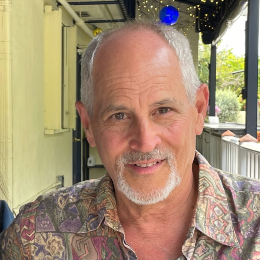Man with gray hair and goatee smiles outdoors in patterned shirt.