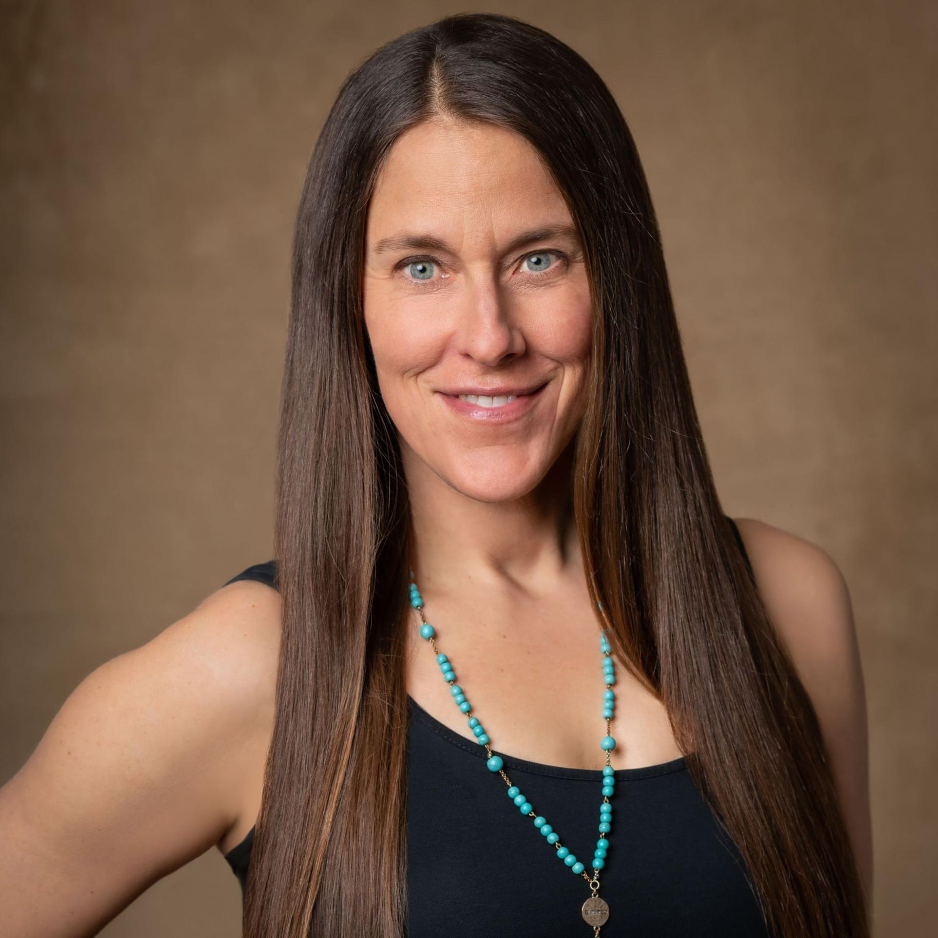 Woman with long brown hair, wearing a black top and turquoise necklace, smiles at the camera against a brown backdrop.