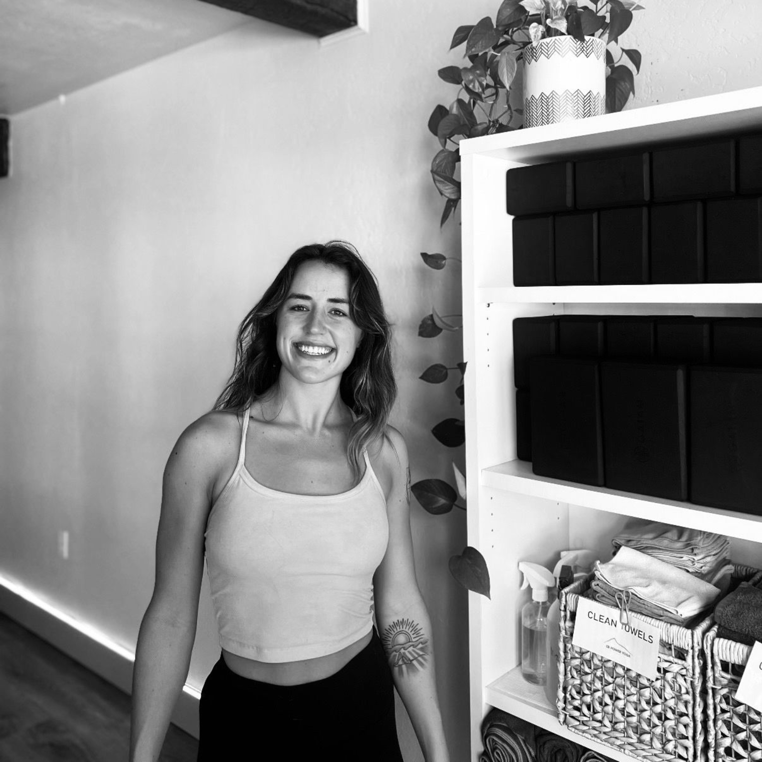 Smiling person in a studio next to shelves with yoga blocks, towels, and plants.