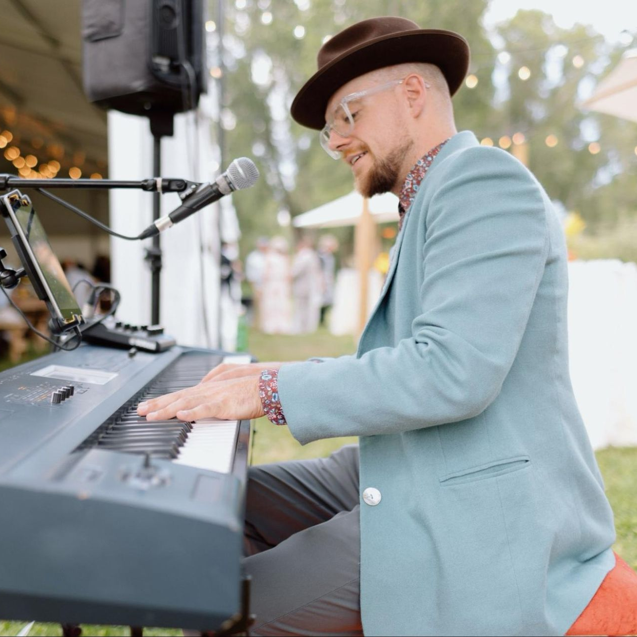 Man playing piano, wearing a hat, blazer, and glasses, outside at an event.