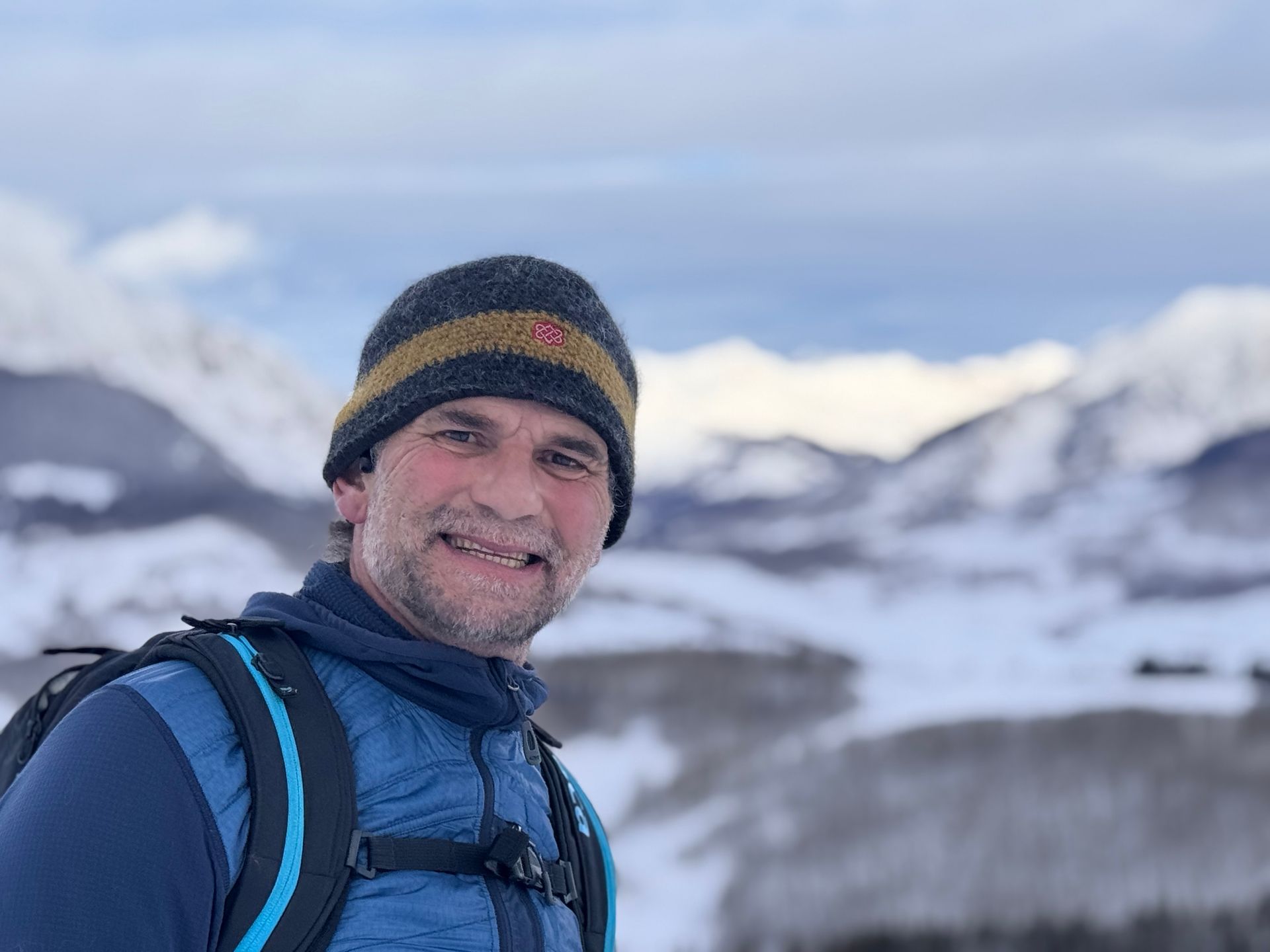 A man wearing a hat and a backpack is standing in front of a snowy mountain.