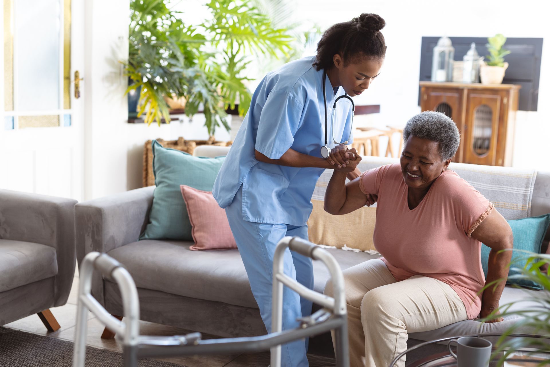 A female nurse helps a female senior standing up from a sofa.