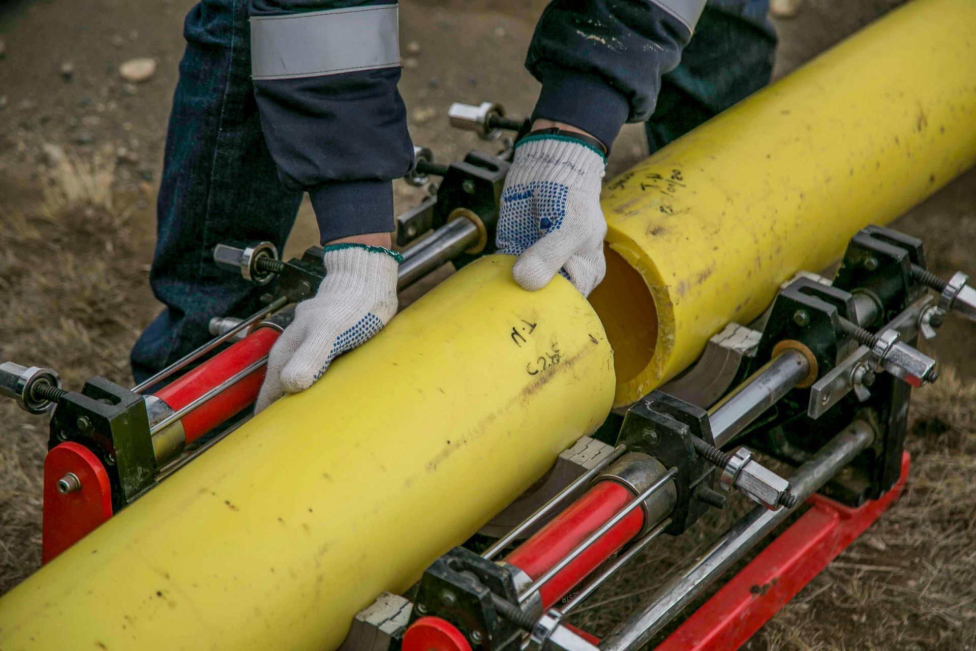 A person in work clothes and gloves uses a mechanical pipe alignment tool to prepare yellow plastic pipes for joining.