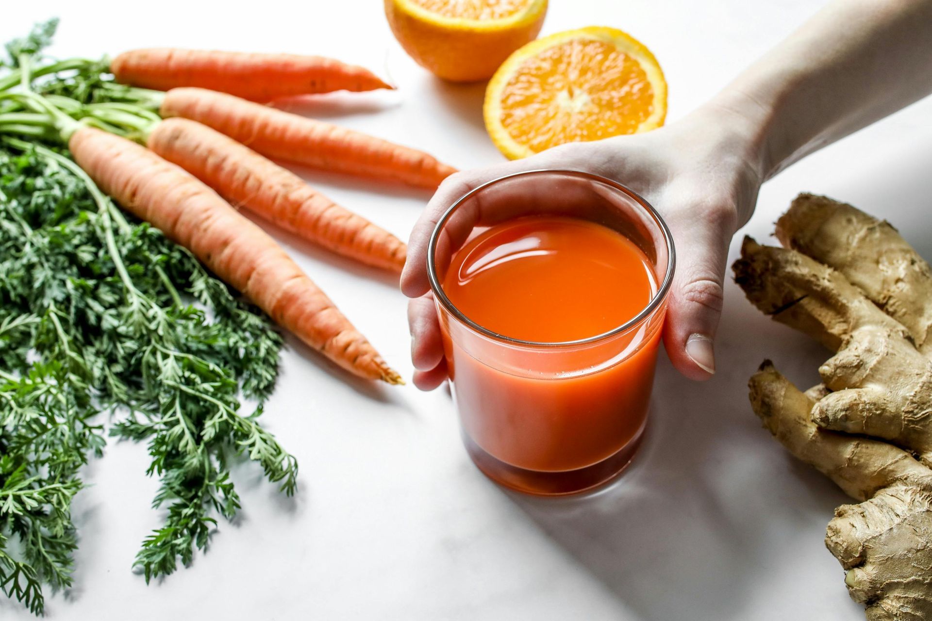 Hand holding a glass of orange vegetable juice with carrots, oranges, ginger, and greens on a white surface.
