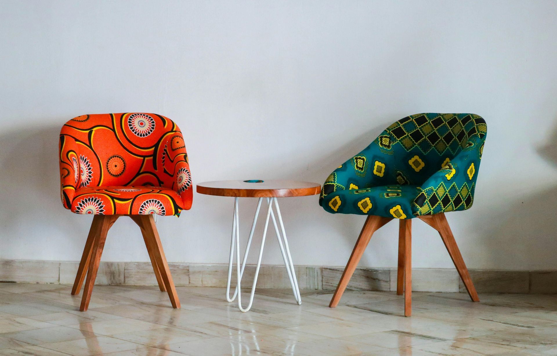 Two colorful upholstered chairs face a small wooden table, against a white wall and tile floor.