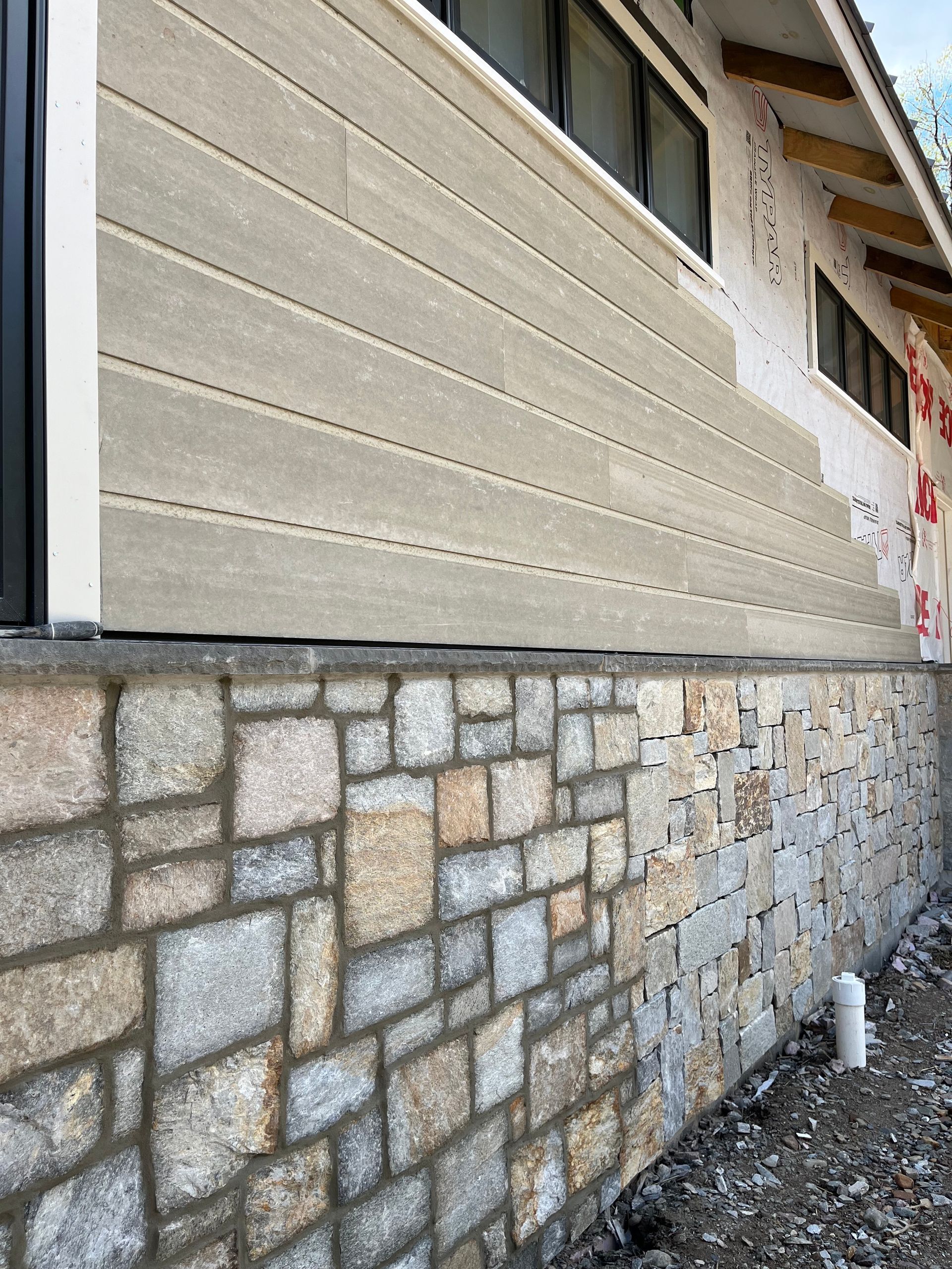 Exterior view of a house with stone foundation and horizontally-laid, light-colored siding. Windows are above.