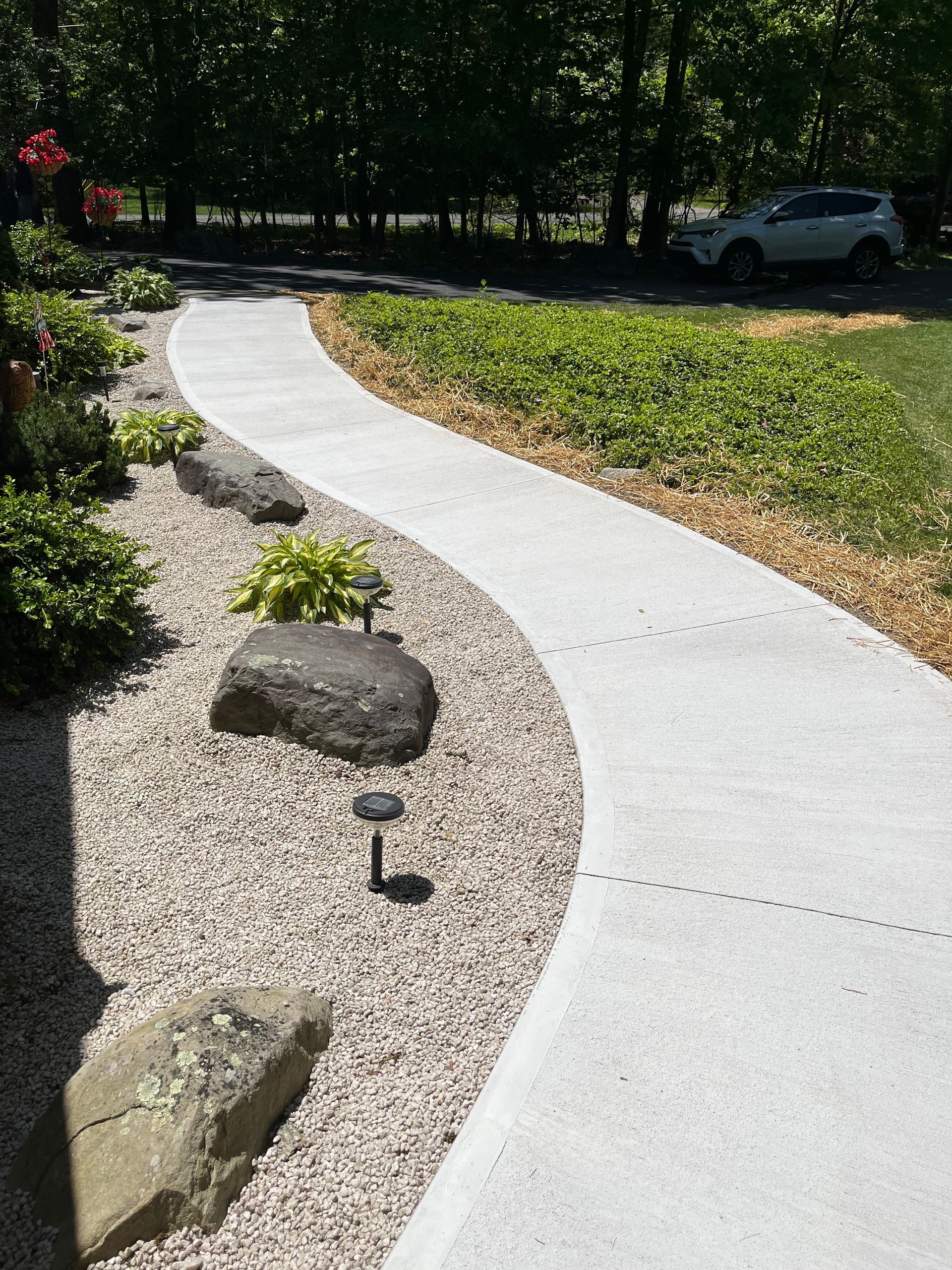 Curving concrete walkway with landscaping, rocks, and gravel. A car sits in the distance.