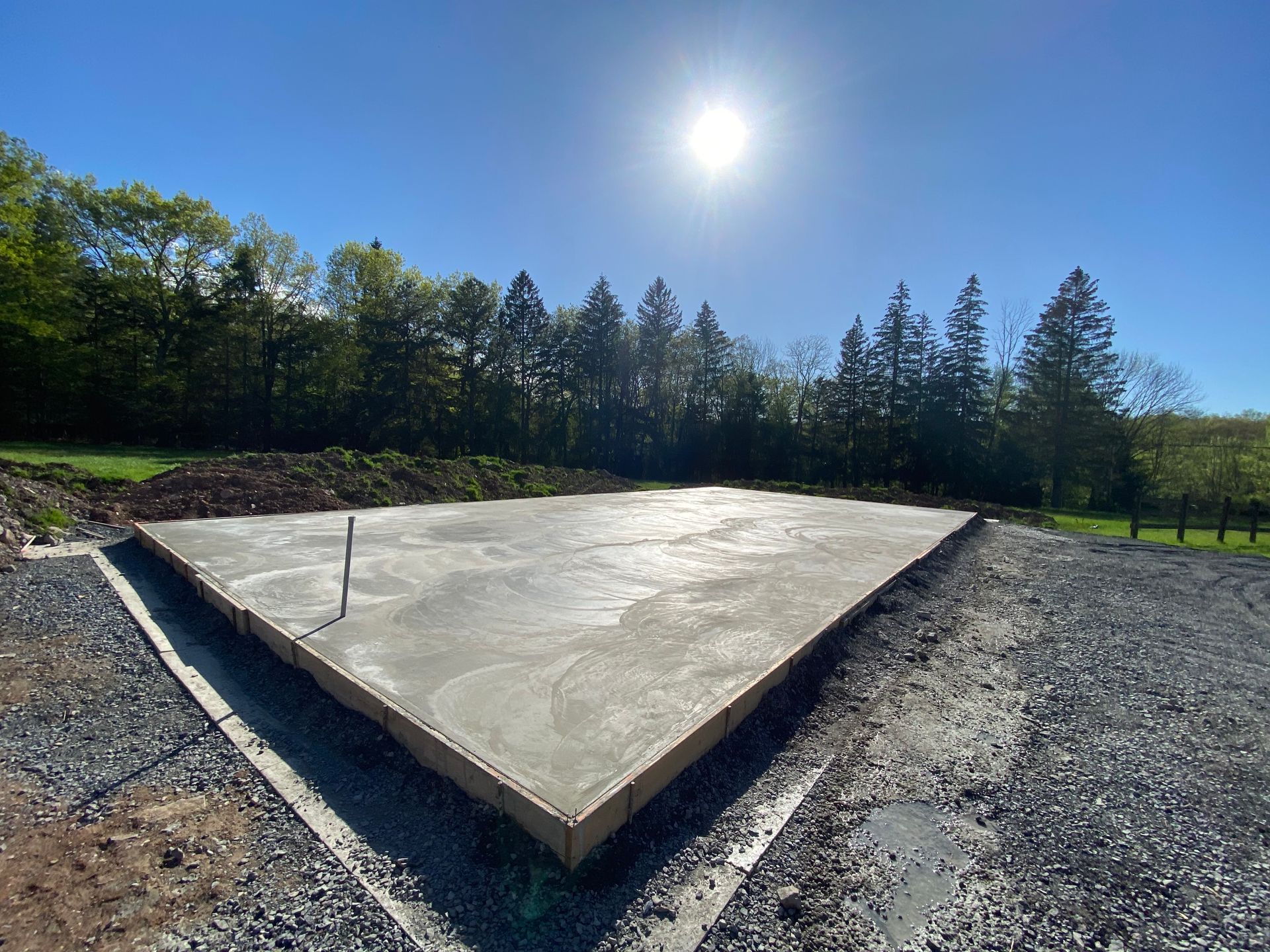 Triangular concrete foundation, freshly poured, bordered by wooden formwork, set on gravel, under a bright sun.