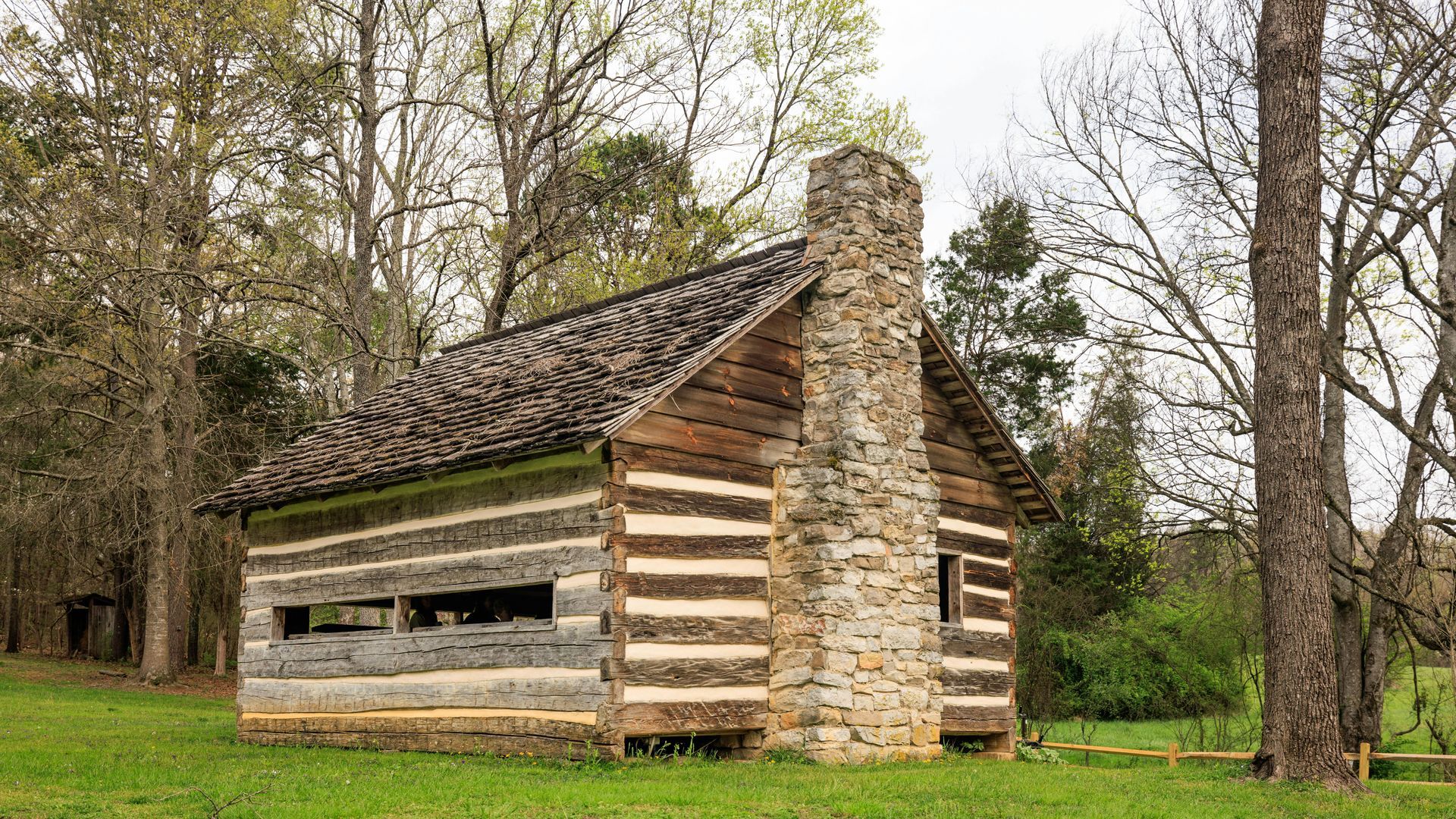A small log cabin with a stone chimney is sitting in the middle of a grassy field.