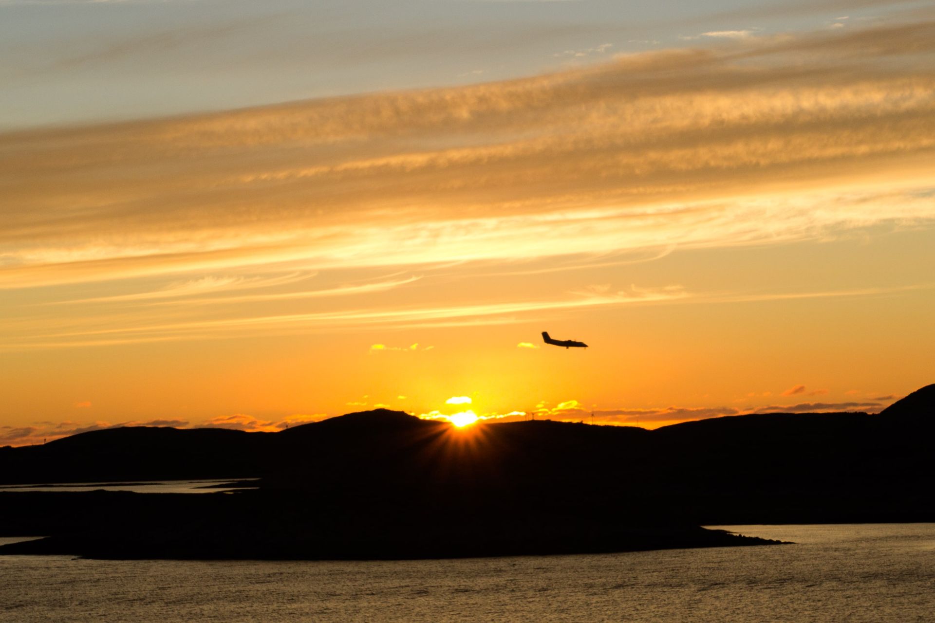 Een vliegtuig vliegt bij zonsondergang over een watermassa en bergen