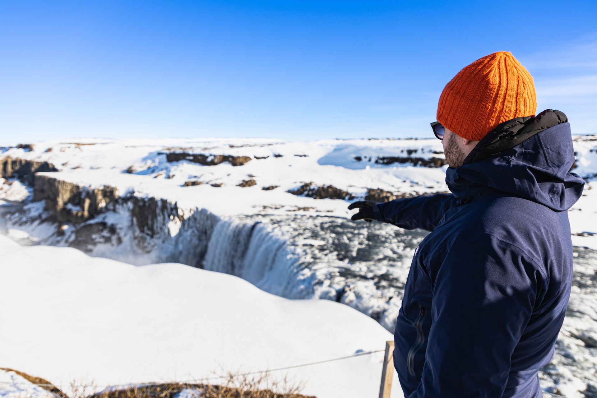 Een man met een oranje hoed wijst naar een waterval in de sneeuw.