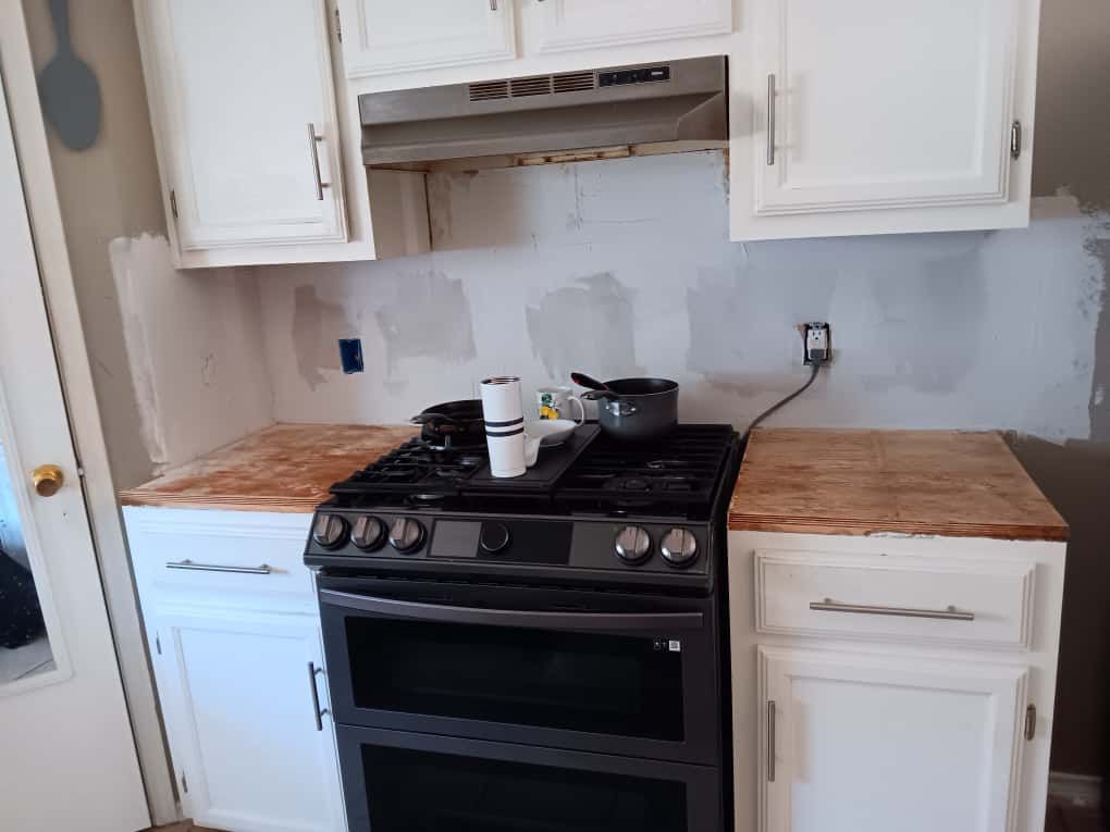 Kitchen under renovation. Gas stove centered between white cabinets with unfinished wood countertops and backsplash.