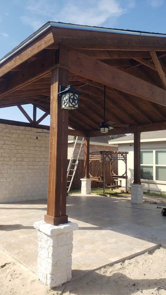 Wooden patio cover with a ceiling fan and light fixture, on stone pillars.