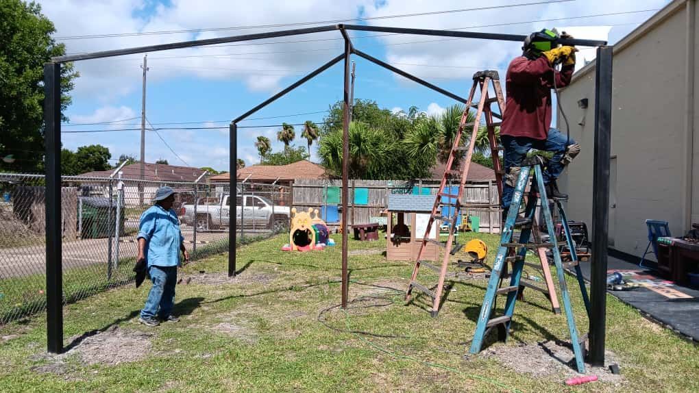Two workers constructing a black metal structure on a grassy lot; one on a ladder welding.