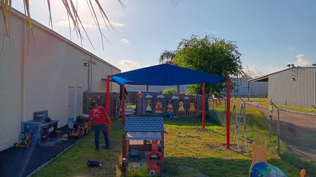 Person standing in a sunny yard with a playground and a blue sunshade.