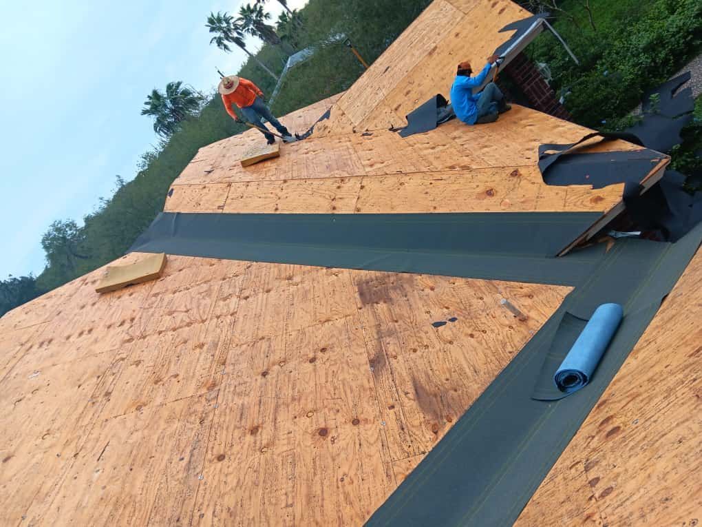 Two roofers install shingles on a partially completed roof, trees in the background.