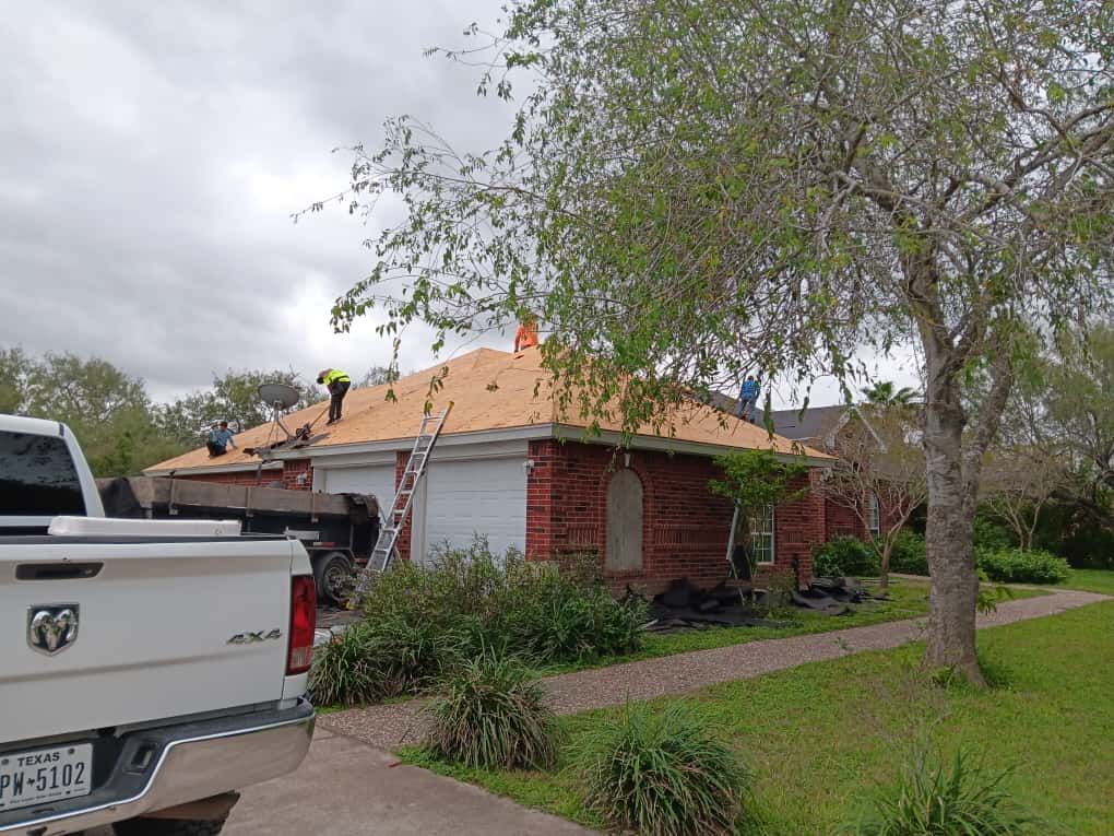 Roofers working on a residential roof; white truck parked nearby, brick house, green grass, cloudy sky.